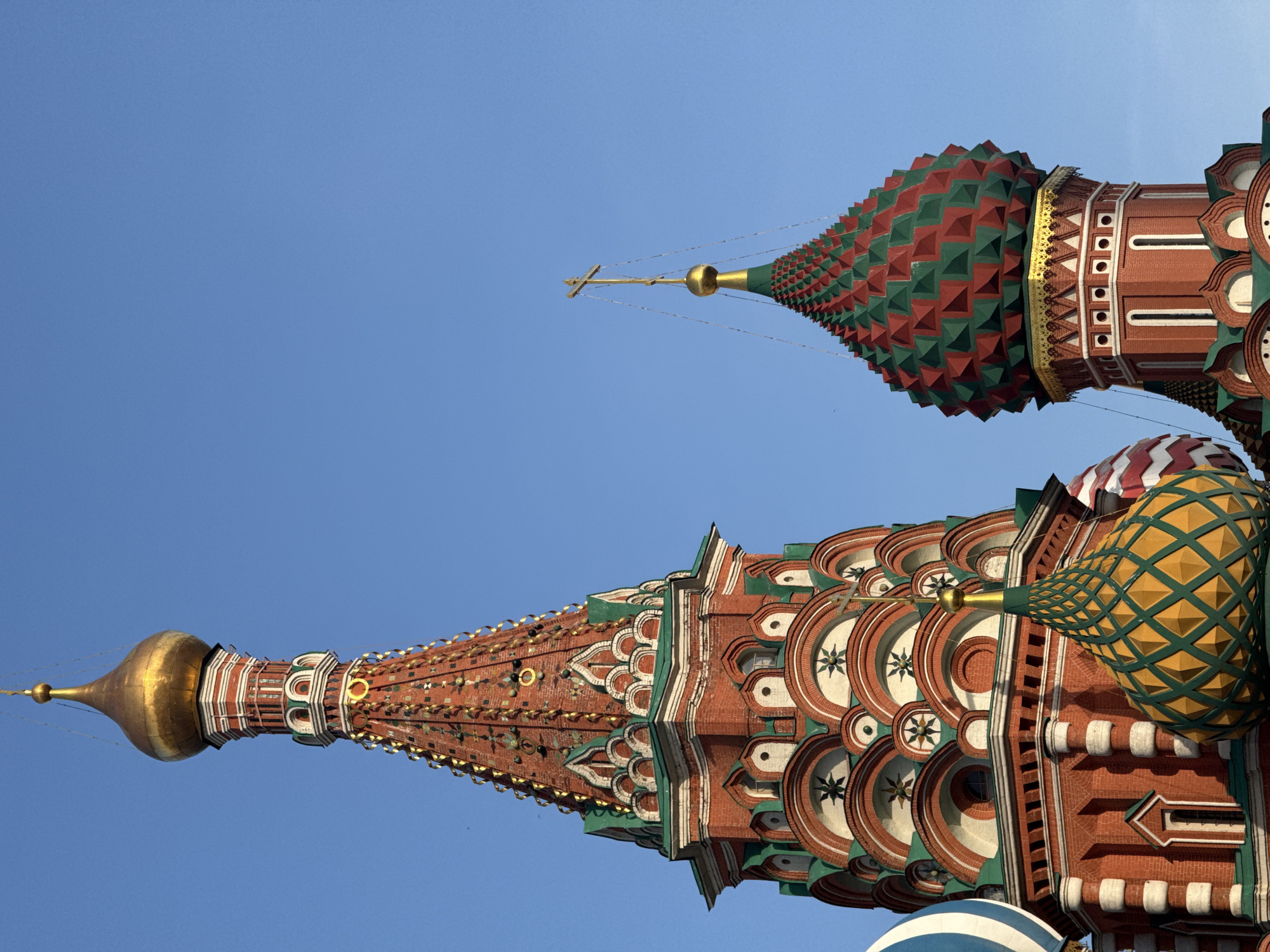 The image is a low-angle view of two intricately decorated domes of St. Basil's Cathedral in Moscow, Russia, set against a clear blue sky. The domes feature a variety of patterns and colors including red, green, yellow, and white. One dome is topped with a gold onion dome and a cross, while the other has a textured surface with green and red pyramid shapes and a cross. The architectural details are ornate, including brickwork and decorative elements.