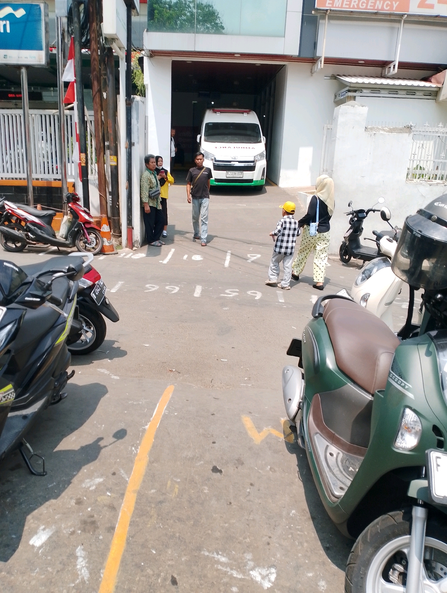 The image depicts an outdoor scene in what appears to be a hospital or clinic parking lot in Bogor, Indonesia. An ambulance is visible in the background, parked near the entrance of a building with "EMERGENCY" signage. Several individuals are present, including two women and a man standing near the entrance, and a woman in a hijab with a child in a yellow hat walking away from the camera. Various motorcycles are parked in the lot, some in the foreground and others further back. The ground is paved and marked with painted lines, suggesting designated parking spaces. The overall environment appears to be a sunny day.