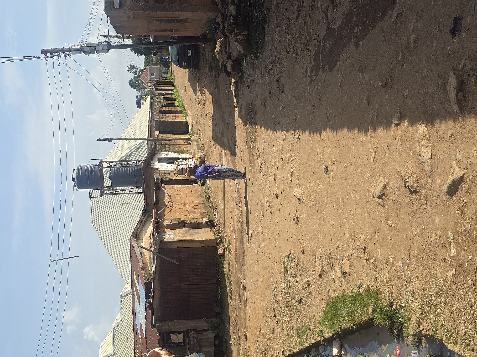 A woman in a blue top and a patterned skirt carries a load of goods on her head as she walks down a dirt road. The road is lined with buildings made of brick and corrugated metal. A water tank sits atop a structure in the background. Power lines crisscross the sky. The scene appears to be in a neighborhood in Jos, Nigeria.