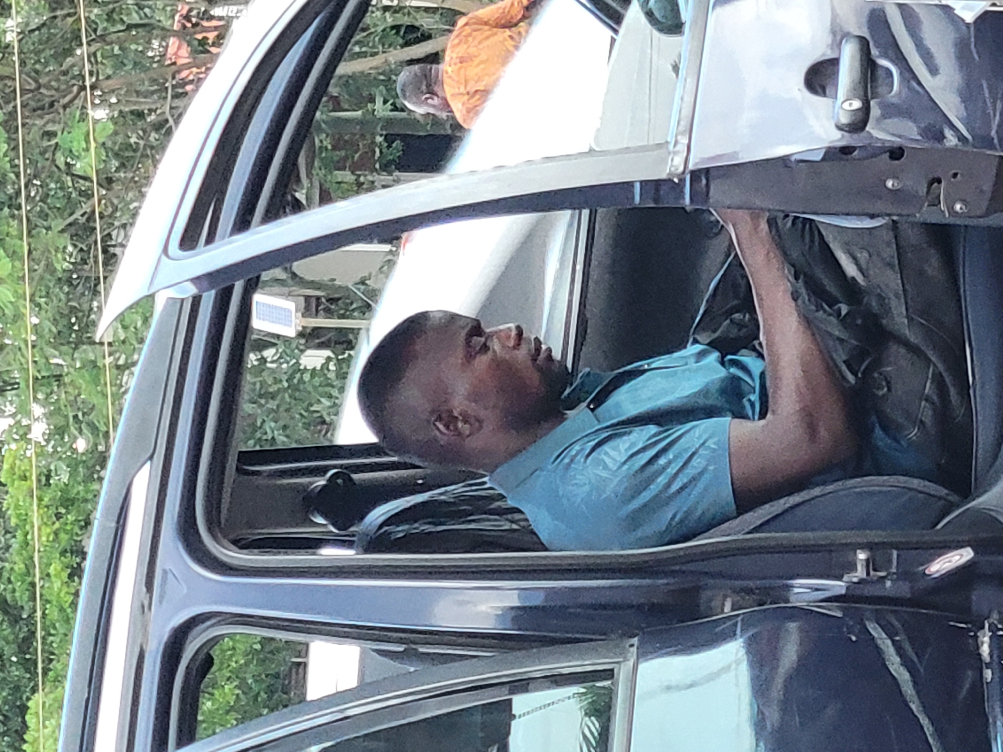 The image shows two individuals and a portion of a dark-colored vehicle, likely an SUV. The primary subject is a man seated in the driver's seat, wearing a teal blue collared shirt. He is looking towards the right of the frame, with his head tilted down and his right arm reaching towards the center console or dashboard area. His expression is not clearly discernible due to the angle.
In the background, another person is visible, wearing an orange garment. This person is further away and partially obscured by the vehicle's door frame and foliage. They appear to be looking away from the camera.
The setting seems to be outdoors, with green foliage and trees visible in the background, suggesting a natural or semi-urban environment. Sunlight is present, indicating it is daytime, though the specific time is unclear. The vehicle's door is open, and the interior is dark. A small white rectangular object, possibly a solar panel or a sensor, is attached to the exterior of the car near the door frame.
There is no visible text in the image. The overall scene suggests a moment of activity related to the vehicle, perhaps an inspection, repair, or adjustment being made by the driver. The focus is on the man in the driver's seat and his interaction with the car's interior.