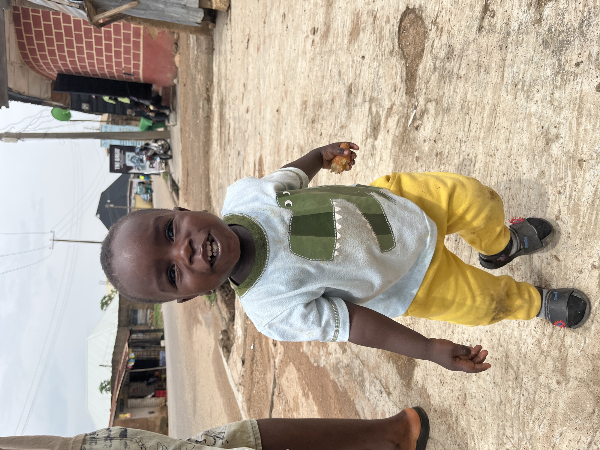 This full-shot image captures a young child standing on a textured concrete surface in what appears to be an urban or semi-urban street setting in Jos, Nigeria. The child, with dark skin and short dark hair, is looking directly at the camera with a wide, open-mouthed smile, revealing a set of white teeth. They are wearing a light blue long-sleeved shirt featuring a green and grey dinosaur design, dirty yellow pants, and black sandals over dark socks. In their right hand, the child holds a piece of light brown food, possibly a fried plantain or similar snack.
The background reveals a bustling street scene under an overcast sky. To the left, there's a building with visible red brickwork, followed by a structure with a dark roof and a green object. Utility poles and power lines crisscross the upper part of the frame. Further down the street, several motorcycles are parked, and various buildings with corrugated metal roofs line the road. Signs are partially visible on some structures; one sign in the mid-ground appears to read "THE GA..." (likely "THE GALLERY"). The ground is cracked and dusty, consistent with a well-used outdoor area. Part of an adult's leg and foot, wearing a black flip-flop, are visible in the bottom left corner of the frame, suggesting another person is nearby. The child's expression conveys joy and happiness. On the child's shirt, the letters "cc" are visible on the sleeve, and faint text can be seen on one of the sandals. The time of day appears to be daytime, with soft, diffused light indicating an overcast sky.