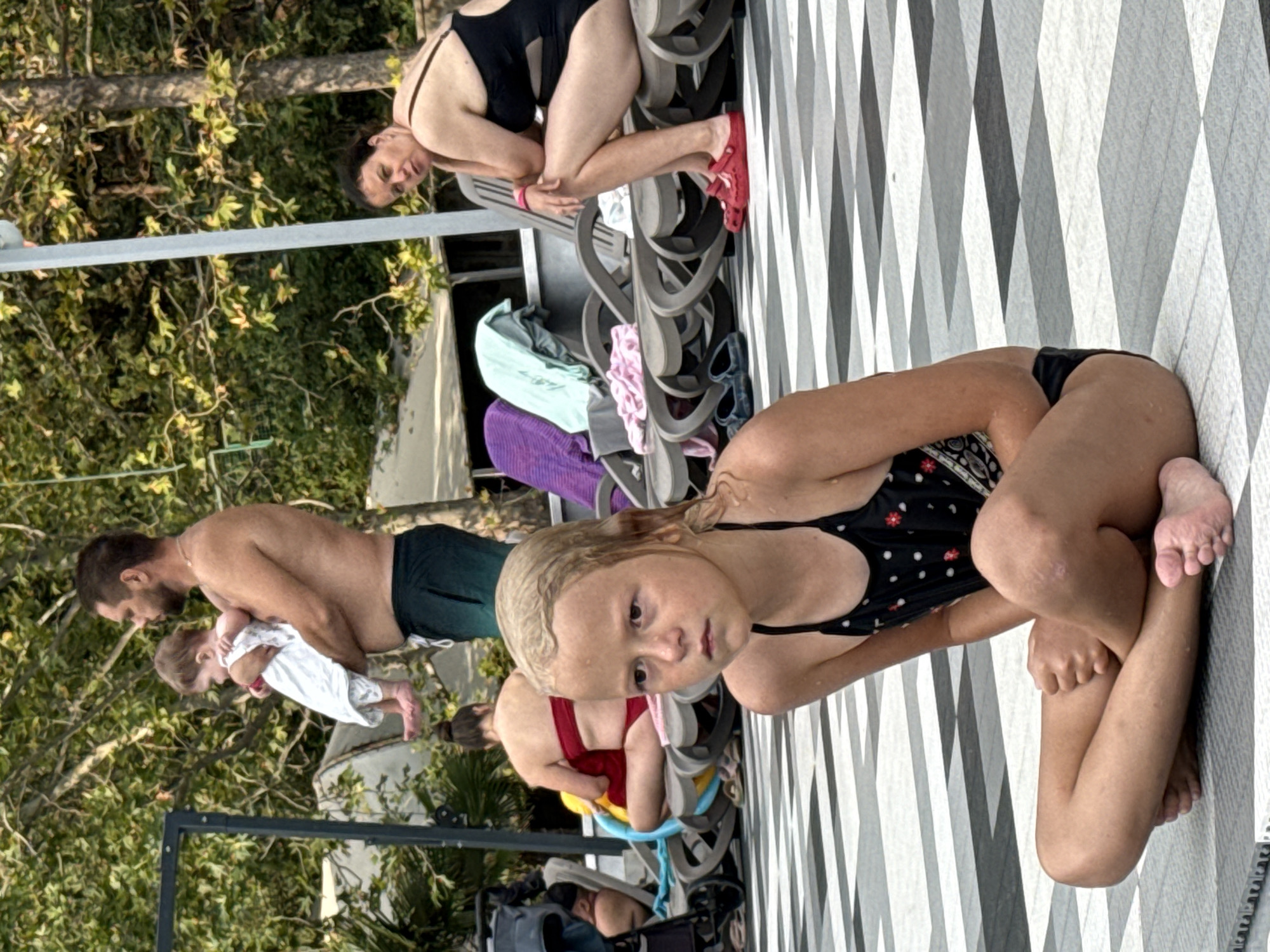 The image shows a group of people by a tiled poolside. In the foreground, a young girl with blonde, wet hair, wearing a black and white polka dot swimsuit, is crouched on the ground. She is looking towards the camera with a neutral expression. Behind her, a man with a beard is holding a baby dressed in white. Further back, a woman in a black swimsuit is sitting on chairs. Several colorful towels are visible near the chairs. The setting appears to be outdoors, with lush green trees and foliage in the background. The ground is covered in a patterned tile design. The lighting suggests it is daytime, likely sunny. There is no visible text in the image.