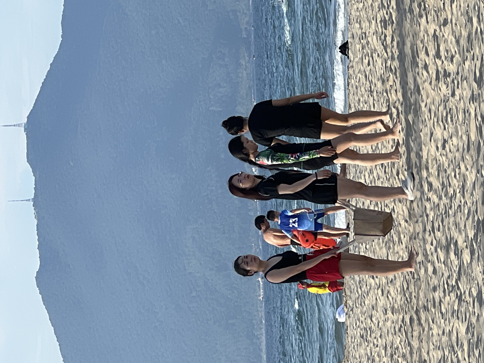 This image depicts a scene on a sandy beach in Đà Nẵng, Vietnam, with the Sơn Trà Peninsula mountain range in the background. The mountain is topped with a distinctive telecommunications tower. In the foreground, several individuals are present on the beach near the ocean.
On the left, a woman wearing a black tank top and red shorts stands facing the camera, holding a light-colored bag with a wooden handle. To her right, a man in red shorts and a yellow life jacket appears to be a lifeguard, looking towards the water. Behind him, a man with his back to the camera, wearing blue swim trunks, is partially obscured. A boy in a blue jersey with the number "23" is also visible. Further to the right, three women are walking away from the camera. One is wearing a black and green patterned swimsuit with black shorts, another is wearing a black outfit with short sleeves and shorts, and the third is wearing a black top and shorts. The ocean is visible behind them, with waves breaking on the shore. The sand is textured and appears to be typical beach sand. A dark object, possibly clothing, lies on the sand near the water's edge on the far right. The sky is bright and hazy, with the mountain range dominating the background.