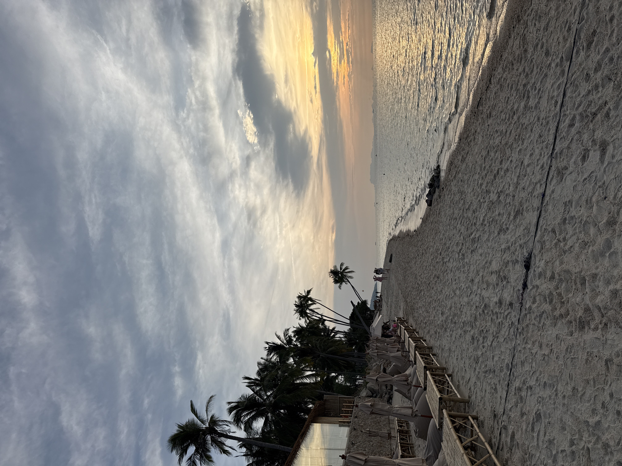 This is a long shot, eye-level, outdoor photograph of a beach scene in Ko Samui District, Thailand, during either sunrise or sunset. The scene is dominated by a sandy beach that stretches into the distance, meeting a calm body of water. The water reflects the warm, golden hues of the sky, suggesting the sun is low on the horizon. The sky itself is filled with a mix of grey and white clouds, with patches of bright yellow and orange indicating the presence of the sun.
Along the left side of the beach, a row of thatched-roof umbrellas and lounge chairs are set up on wooden platforms. Behind these, a modern-looking building with large glass windows and a thatched roof is visible, flanked by numerous palm trees that lean towards the sea. A few people are visible on the beach further down, walking or standing. In the distance, the silhouettes of islands or land masses can be seen against the horizon, with a few small boats dotting the water. The sand itself is textured with footprints, indicating recent activity. A black cable or rope runs across the sand in the foreground.