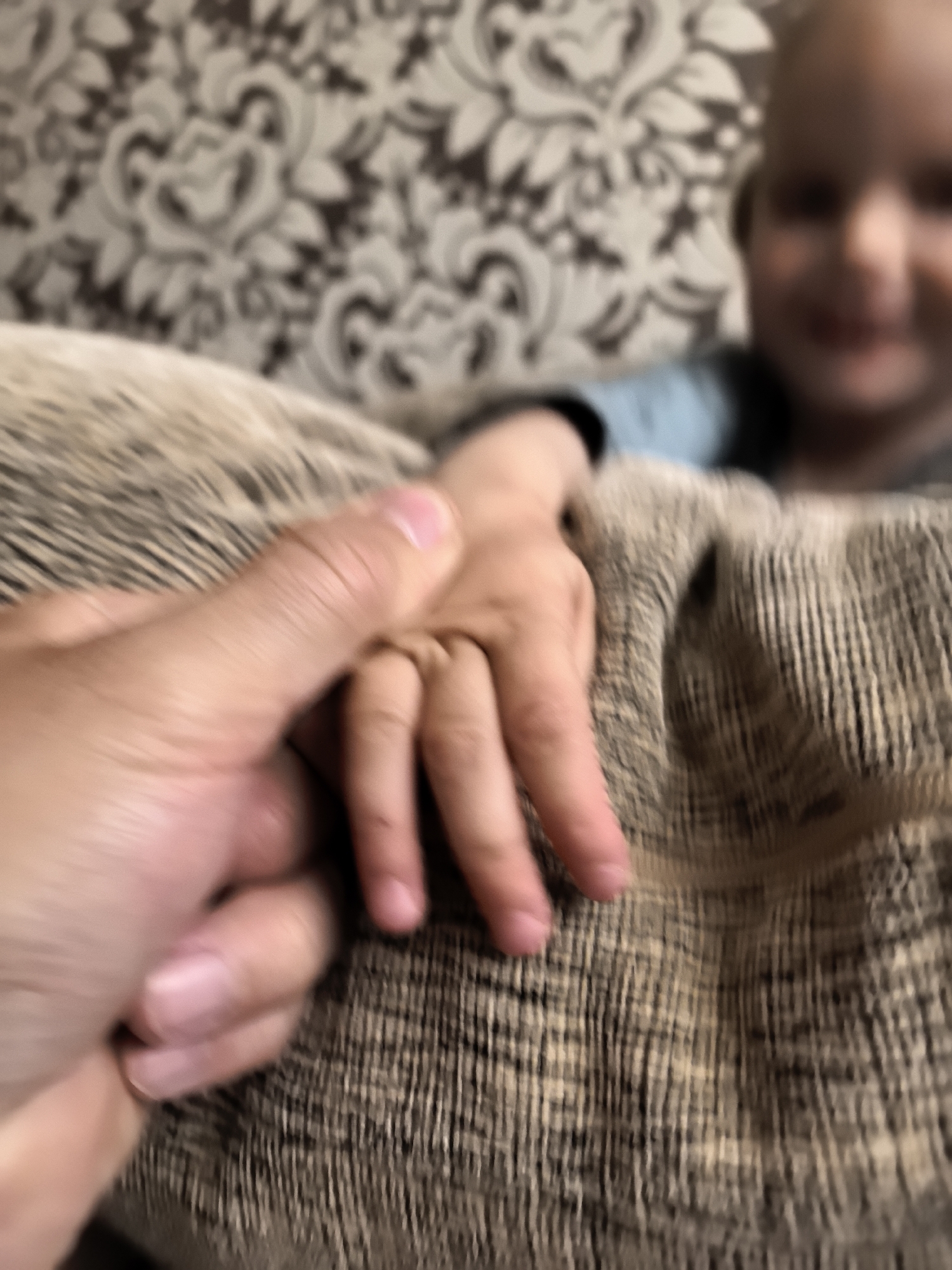 An adult's hand clasped around a child's hand is shown in extreme close-up. The adult's hand is in the foreground, with the child's hand visible in the center. The background is out of focus and appears to be a patterned fabric. A child's face is partially visible in the upper right corner, blurred. The lighting is soft and warm.