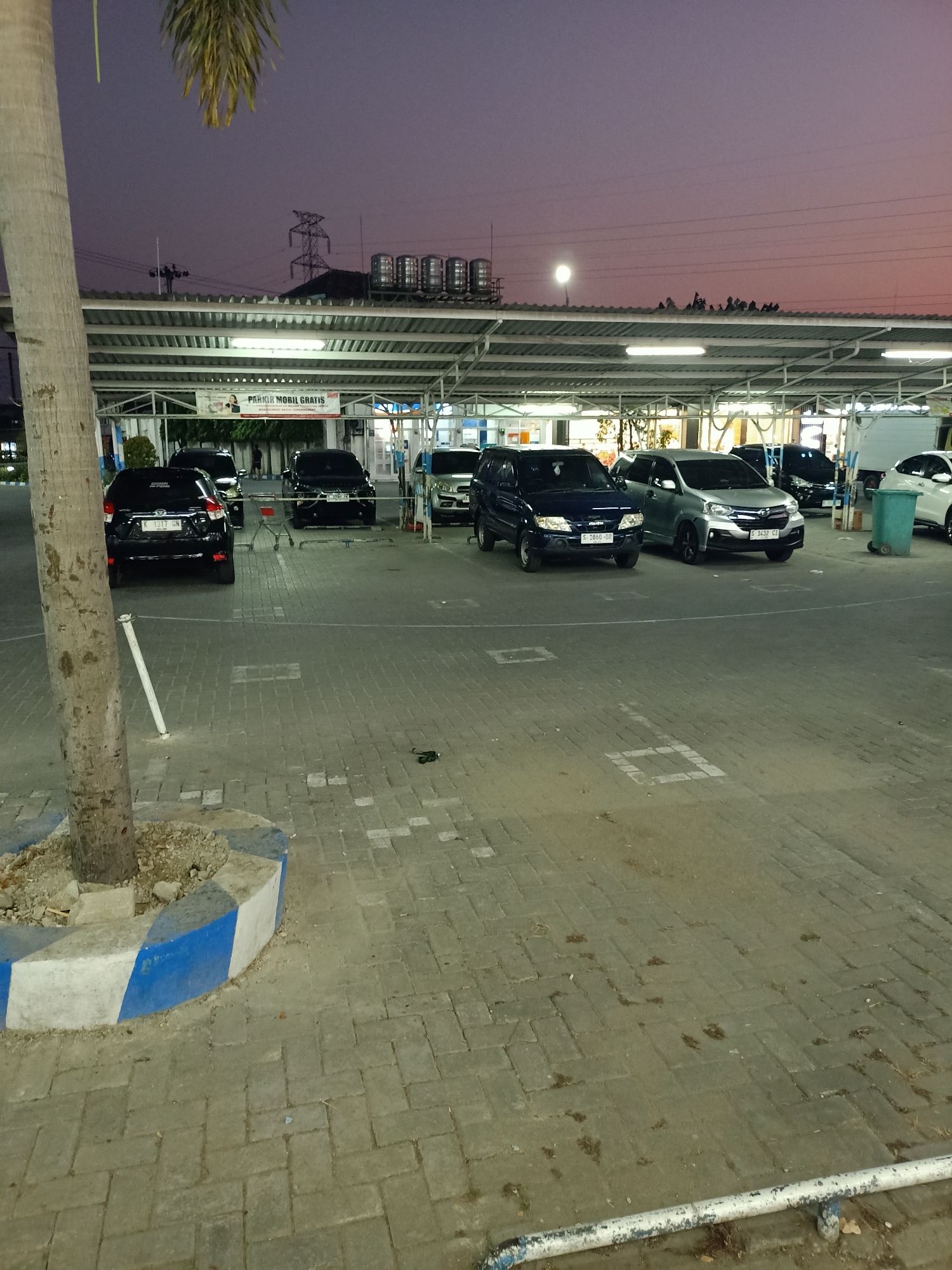This image shows a parking lot at dusk in Mernung, Indonesia. Several cars are parked under a covered structure, including black SUVs and a silver minivan. A palm tree stands to the left of the frame, and a sign above the parking area reads "PARKIR MOBIL GRATIS" (Free Car Parking). The ground is paved with interlocking bricks. In the background, power lines and what appear to be water tanks are visible on top of a building. The sky is a deep purple, indicating the transition from day to night.