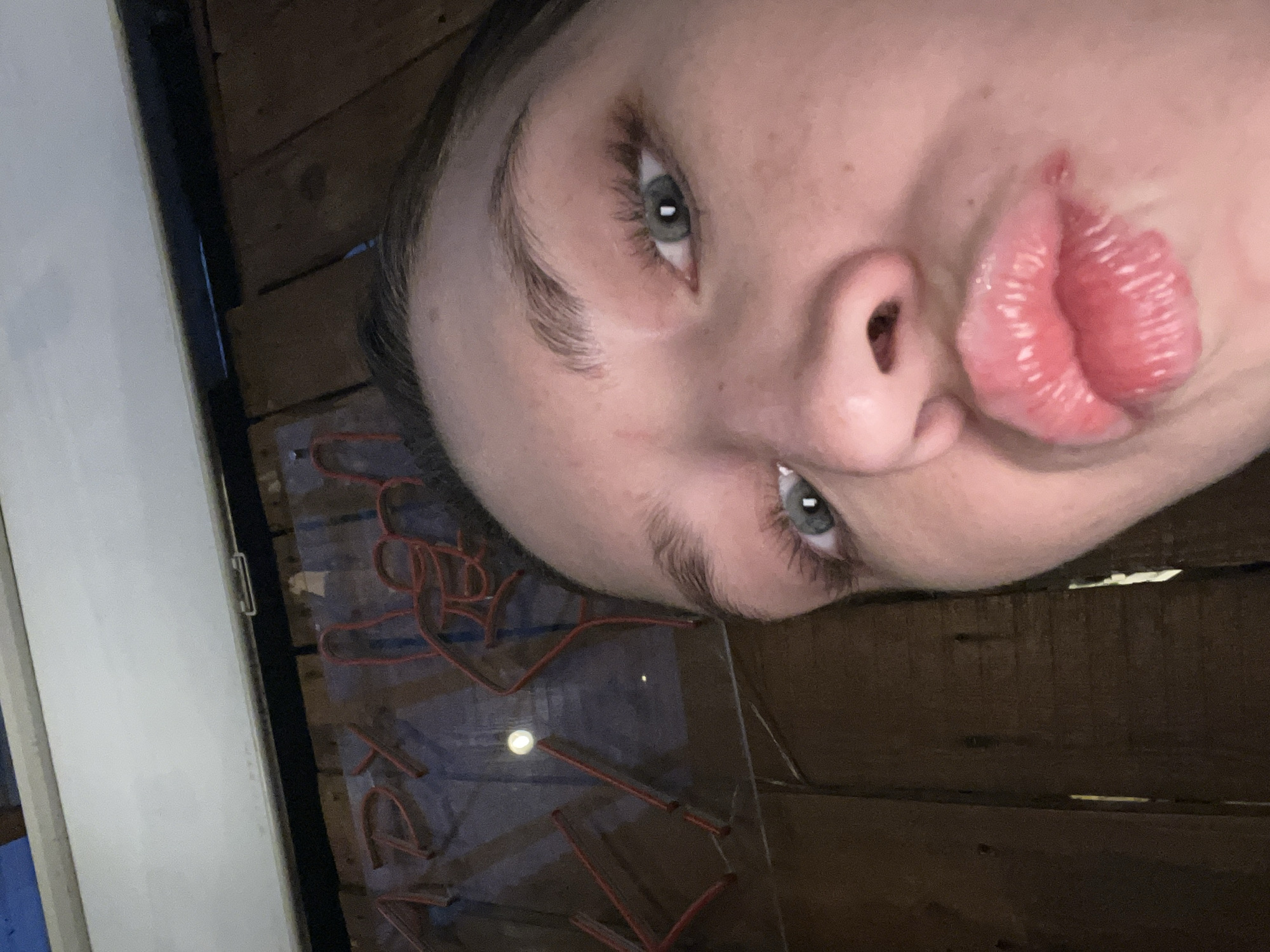 A person's face is angled towards the camera with a pronounced pout. The person has light skin, light-colored eyes, and visible eyebrow hairs. A red neon sign is partially visible, positioned below the person's face. Wooden planks form the background. No indication of location is apparent in the image.