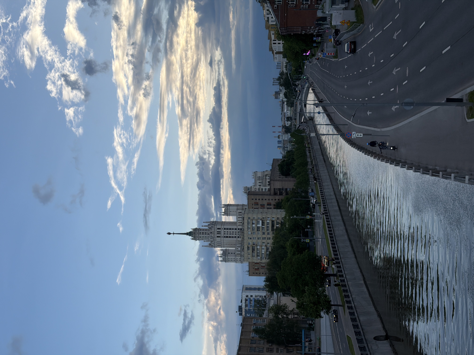 The image captures an urban cityscape under a partly cloudy sky. The primary focus is a tall, Soviet-era skyscraper, likely one of the Seven Sisters in Moscow, Russia. The building features a spire and multiple tiered sections, typical of Stalinist architecture.
To the right of the building, a wide avenue runs parallel to a river. Vehicles, including cars and a motorcycle, are visible on the road. The riverbank is lined with a concrete barrier, and trees are planted along the adjacent sidewalk. The sky is partly covered with clouds, some illuminated by the setting sun, suggesting it might be early evening. Buildings and other urban structures are visible in the background.