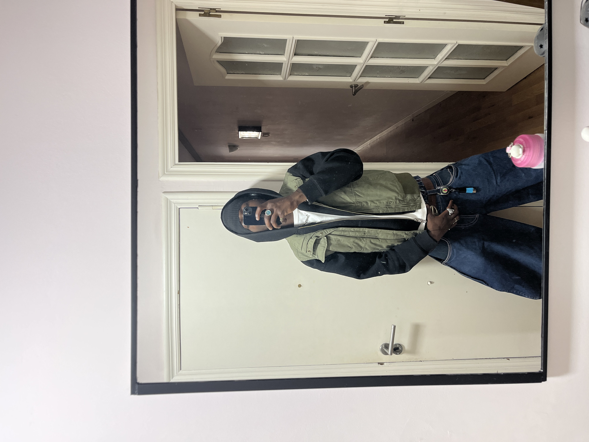 A mirror reflects a person wearing a black hooded jacket and olive-green vest over a white shirt, with dark blue jeans. The reflection shows the person holding a phone, presumably taking the picture. In the background, a door with rectangular panels is visible, above a hallway. A pink water bottle sits atop the right-hand side of the reflection and a doorknob is present in the reflection. The image is contained within a black-framed mirror. The location is likely indoors, with a neutral wall. There is no indication of the location being in the city of Barcelona, Spain.
