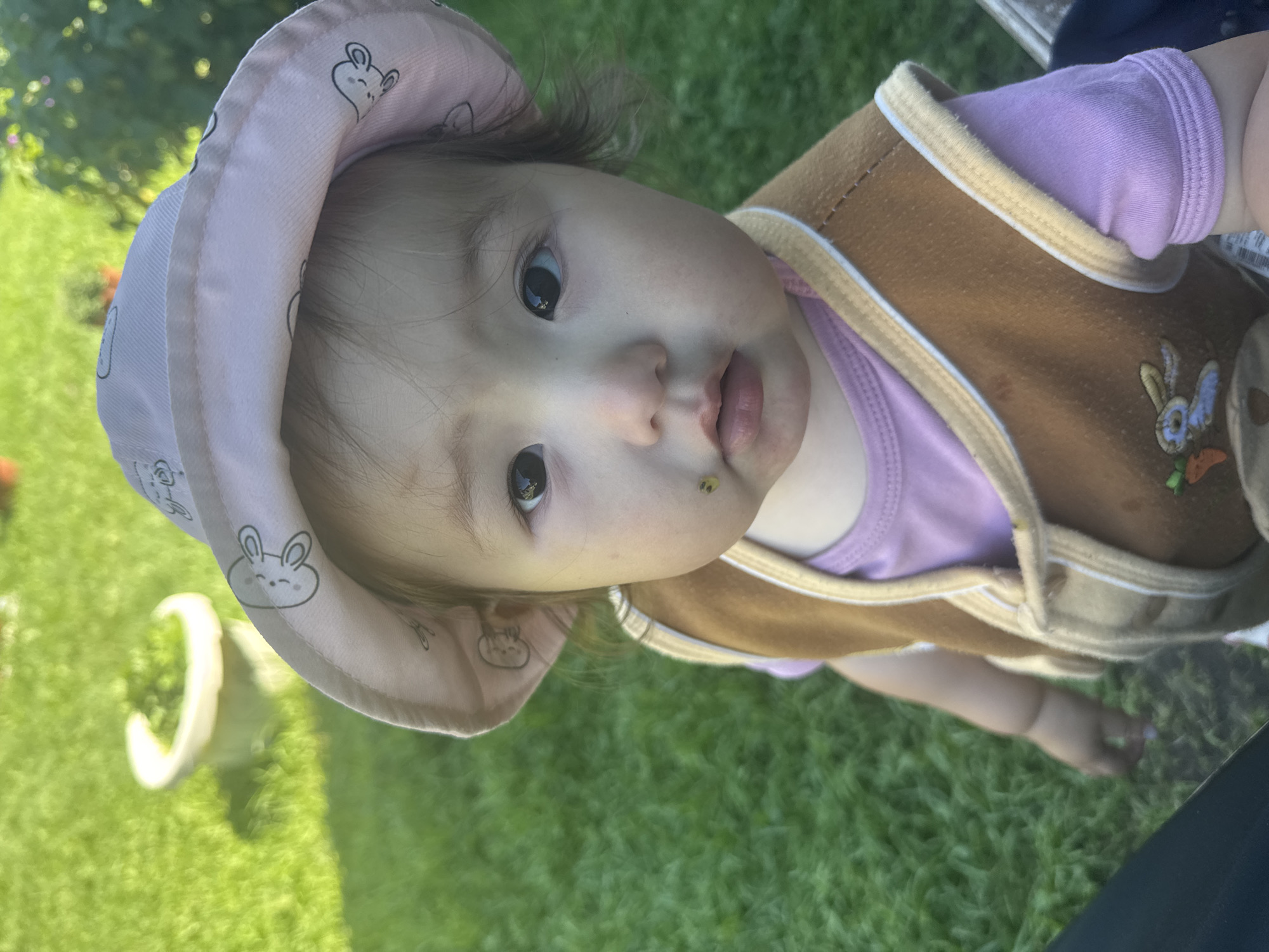 A close-up, eye-level shot shows a young child wearing a pink bucket hat with rabbit patterns and a brown vest over a lavender t-shirt. The child has dark eyes and a small smudge of green on their cheek near their mouth. The background is out of focus, showing green grass and a light-colored pot with greenery. The scene appears to be outdoors on a sunny day. There is no information in the image to indicate a location in the city of Yabogan, Russia.