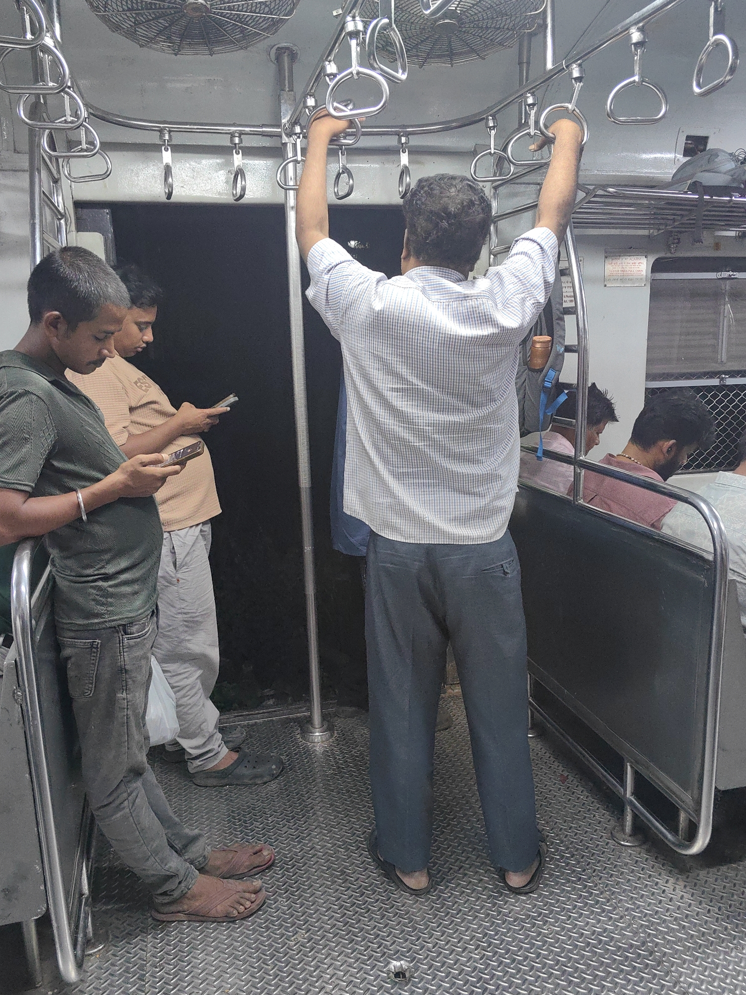 This is a photograph taken inside a crowded Mumbai local train. The image shows several passengers standing and holding onto the overhead bars for support. In the foreground, a young man in a green t-shirt and grey jeans is looking at his phone. Next to him, another man in a beige t-shirt and light grey pants is also engrossed in his mobile device. A third man, wearing a checkered shirt and dark trousers, is standing further back, holding onto a grab handle. Other passengers are visible in the background, some seated and others standing. The floor of the train is made of patterned metal. The lighting suggests it is either daytime with limited light penetration or evening. The overall atmosphere depicts the typical rush of a Mumbai local train commute.