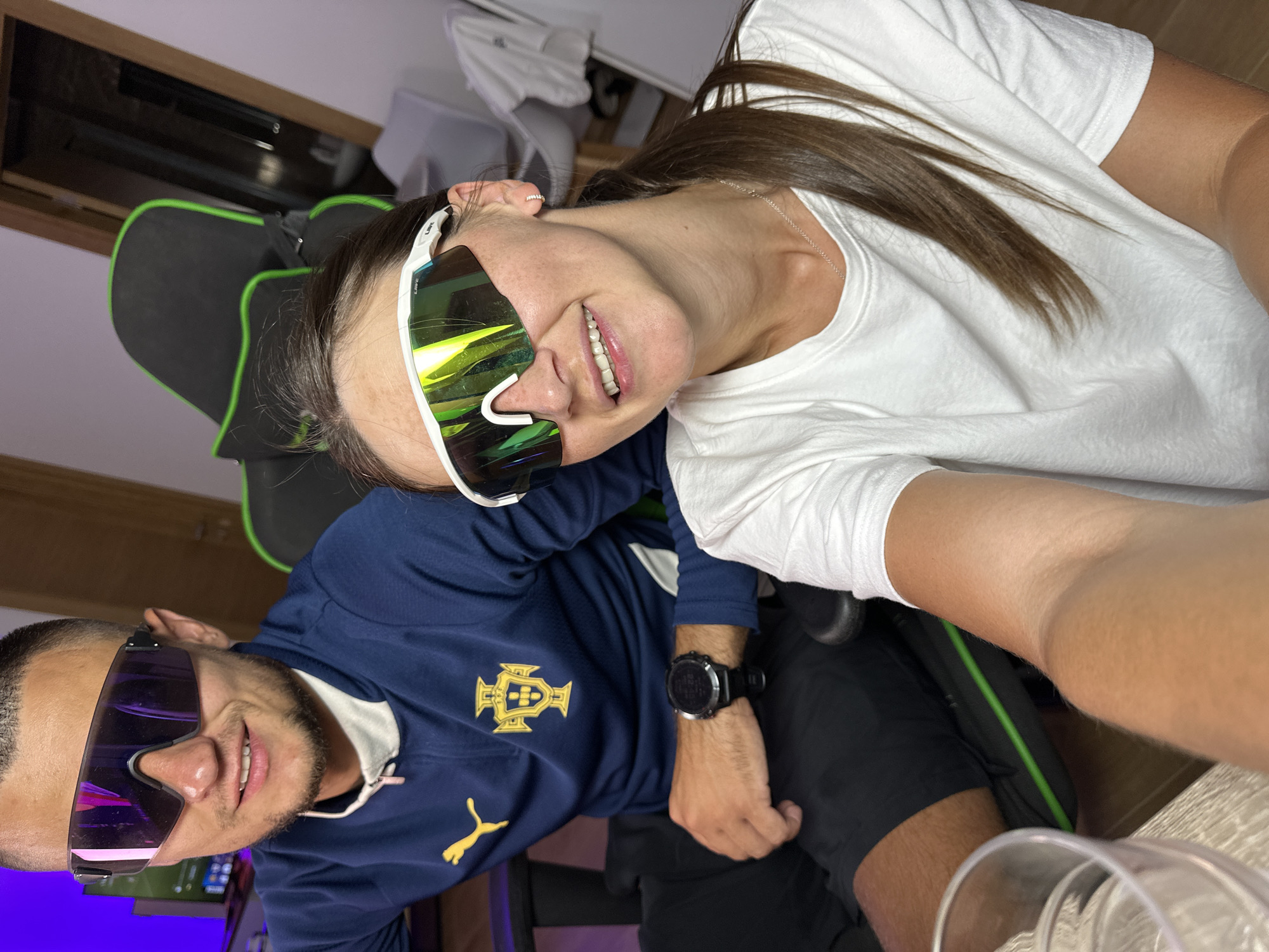 The image shows a selfie taken by two individuals. The person in the foreground is a woman with long brown hair, wearing a white t-shirt and large, white-framed, iridescent sunglasses. She is smiling and looking towards the camera. Behind her, a man is visible, also wearing sunglasses, these with darker, purplish lenses. He is wearing a navy blue long-sleeved shirt with a white collar and a yellow emblem on the left chest. He has a watch on his left wrist. Both individuals appear to be sitting in gaming chairs, suggested by the black and green upholstery visible in the background. The scene is indoors, with a wooden surface in the foreground, possibly a desk, and a hint of a computer monitor on the left. The lighting suggests it could be daytime or well-lit indoors. The mood seems casual and friendly. The emblem on the man's shirt is yellow and depicts a crest with a shield and a stylized symbol above it, and a Puma logo is also visible on his shirt. The sunglasses worn by both individuals are sporty and oversized.