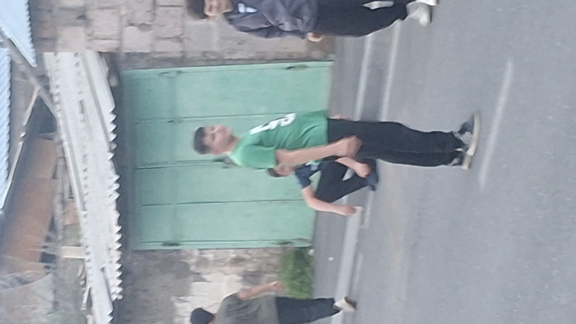 The image shows a street scene in Gyumri, Armenia, with several young people visible. In the foreground, a person wearing a green t-shirt with white lettering and black pants is standing and facing to the right. To the left, another person in a dark t-shirt and black pants is walking away from the camera. In the middle ground, a person is crouched down wearing a blue t-shirt and black pants. Behind them, a light green gate is visible, along with a stone-built structure. The ground is paved and appears to be asphalt. Another person in a grey hoodie and black pants is standing on the right side of the image. The overall lighting suggests it is daytime.