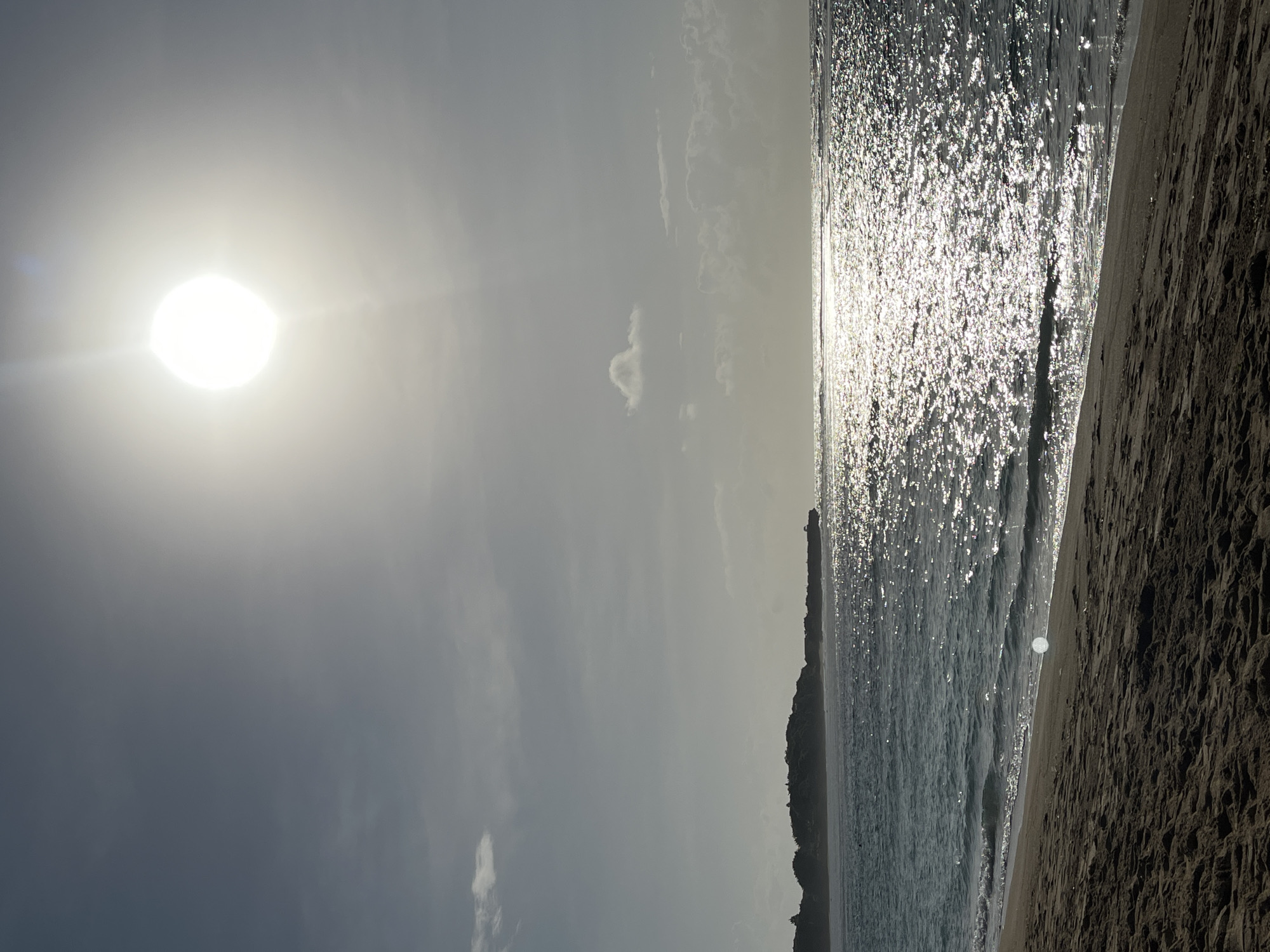 This is a photograph of a beach scene in Kuta Selatan, Indonesia. The image is oriented vertically. The sun is prominently displayed in the upper left of the frame. The ocean spans the right side of the picture, reflecting the sun's glare. The beach is located along the bottom edge. A line of vegetation is visible along the shoreline, separating the beach from inland areas. There is no evidence of any people in the image.