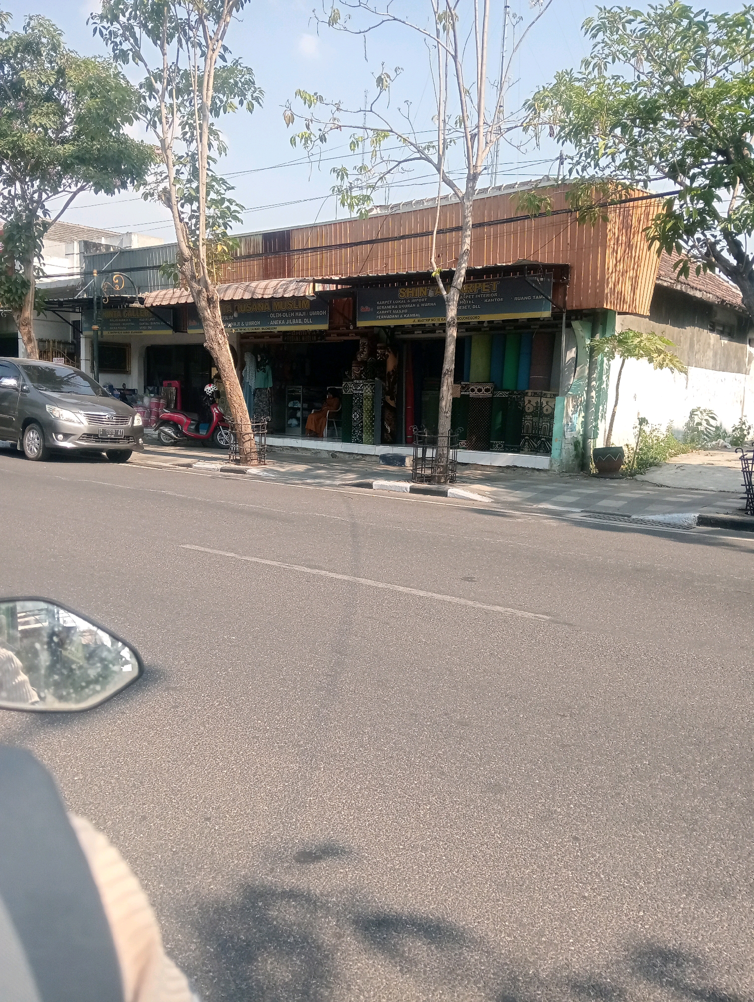 The image shows a street in Bojonegoro, Indonesia, with a storefront on the right side. The storefront displays a variety of textiles, including brightly colored fabrics and patterned cloths, arranged on shelves. A sign above the storefront reads "TINCA MUSLIM" and "SHIN AZZA CARPET". There are several trees in front of the building. A grey SUV is parked on the left side of the street, and a red scooter is parked near the storefront. The road is paved, and there is a sidewalk in front of the building. A motorcycle's rearview mirror is visible in the lower left corner, indicating the photo was taken from a motorcycle. The sky is clear and blue.