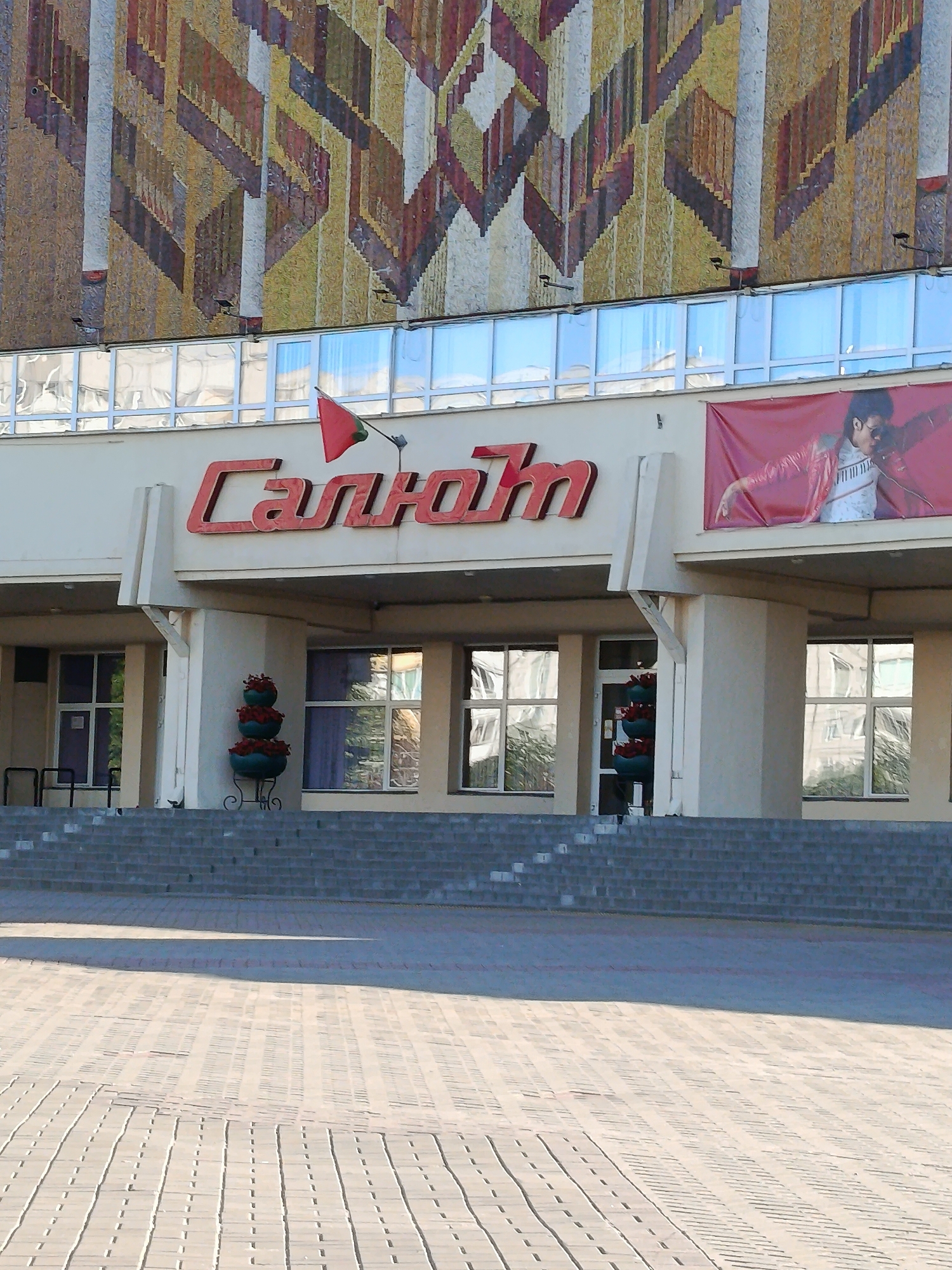 The image shows the facade of a building with the Cyrillic lettering "Салют" in red neon lights above the entrance. A red banner featuring a person in a red jacket and sunglasses is displayed to the right of the lettering. The building has large windows and is accessed by a set of stairs. Planters with red flowers are visible near the entrance. The building is located in Minsk, Belarus.