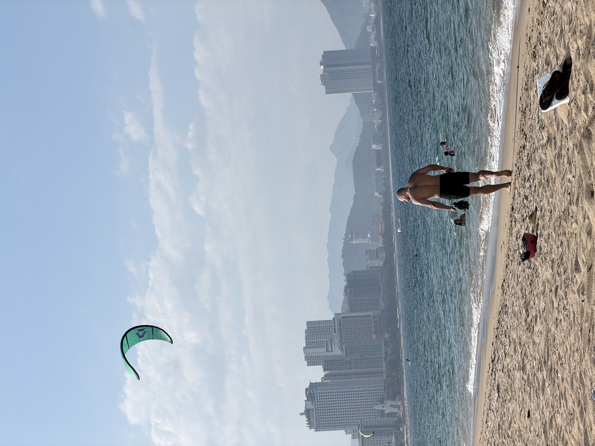 The image depicts a daytime scene on a sandy beach in Nha Trang Ward, Vietnam, with a city skyline and mountains in the background. In the foreground, a shirtless adult male wearing black swim shorts walks barefoot towards the ocean, holding a dark green item in his left hand. To his right on the sand, a pair of dark flip-flops rests on a white cloth, and further right, a pink bucket or container is visible. The sandy beach shows various textures and minor debris.
The turquoise-blue ocean features gentle waves breaking on the shore. Several individuals are in the water; two heads are clearly visible to the left of the walking man, and another head is visible further right. A faint silhouette of a person on a board appears further out in the water, likely kitesurfing. High in the sky, a green and black kitesurfing kite is prominently displayed against a light blue sky with scattered white clouds. Another kite is faintly visible closer to the horizon on the left.
The background presents a dense urban landscape of Nha Trang Ward, characterized by numerous high-rise buildings, predominantly white or light-colored, lining the coast. A distinctive white, pointed structure resembling a church or monument is visible near the waterline to the left. Behind the city, a range of large, hazy blue-grey mountains extends across the horizon, partially obscured by atmospheric conditions. The overall lighting suggests a sunny day.