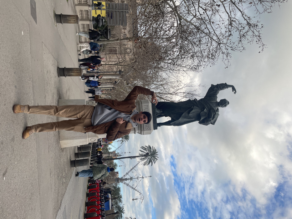 The image displays a young man posing playfully next to a large statue in what appears to be an outdoor public square or promenade. The photo is rotated 90 degrees clockwise.
In the foreground, a young man stands upright, facing slightly towards the camera. He is wearing a light gray hooded sweatshirt under a brown or tan jacket, light brown/beige pants, and matching lace-up boots. His left arm is bent, with his hand raised and open, seemingly mimicking the pose of the statue beside him. His right arm is also bent, with his hand near his chest. His mouth is open, suggesting he is either speaking, laughing, or expressing excitement for the photo.
To the man's immediate right is a prominent statue resting on a light-colored stone pedestal. The statue is dark-colored, likely bronze, and depicts a male figure in period attire with one arm raised in a gesturing or pointing manner. The base of the pedestal clearly shows the text "PAU CLARIS".
The background reveals a wide paved area with several bollard-like structures extending into the distance. Numerous other people are visible further down the path, some walking, others standing, adding to the sense of a busy public space. Beyond the paved area, there are bare deciduous trees, suggesting a late autumn or winter season. Behind the trees, grand architectural buildings with detailed facades can be seen.
In the mid-ground and background, particularly to the right of the statue, several palm trees are visible, and in the lower right corner (if correctly oriented), there are rows of red bicycles, likely part of a public bike-sharing program.
The sky is partially cloudy, with a mix of gray clouds and patches of clear blue sky. The lighting suggests it is daytime, possibly late morning or early afternoon. Based on the visible text "PAU CLARIS" on the statue, the location is likely Barcelona, Spain, where a monument to Pau Claris i Casademunt is situated.