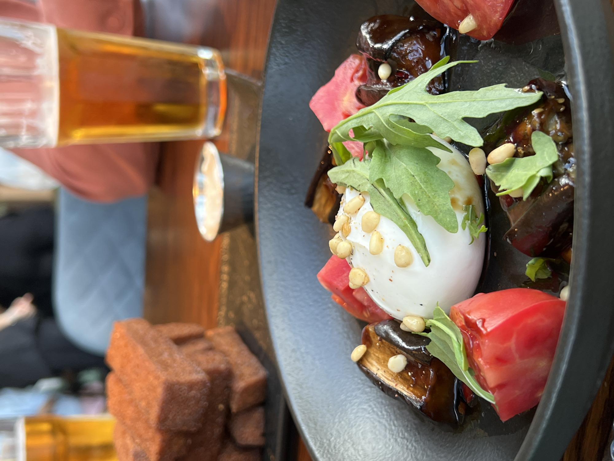 An overhead close-up shot shows a dark gray, circular plate filled with food. At the center of the plate is a large, white, spherical object that appears to be burrata cheese, topped with a few green leaves and scattered pine nuts. Surrounding the burrata are chunks of roasted eggplant, glistening with a dark sauce, and wedges of bright red tomato. Arugula leaves are scattered throughout the dish, adding a touch of green. To the left of the plate, a stack of small, rectangular pieces of dark bread, possibly toast or croutons, is visible. In the background, a glass of amber liquid, likely beer, is on the right, with another glass of beer and blurred figures of people seated at a table. The setting appears to be a restaurant or cafe, with a wooden table surface. The scene is set in Khimki, Russia.