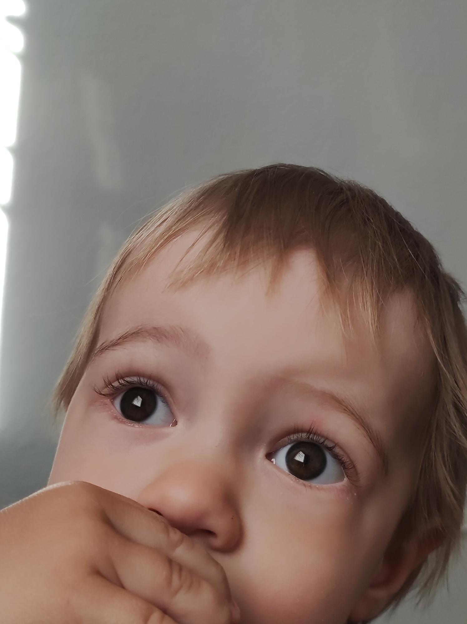 A close-up shot of a young child with light brown hair and dark brown eyes looking upwards and slightly to the left. The child's hand is partially covering their mouth. The background is a blurry, light gray surface with indistinct bright vertical lines on the left side.
There is no verifiable information to confirm the location of the image as being in Chaikovsky, Russia. The image provides no contextual clues about the location.