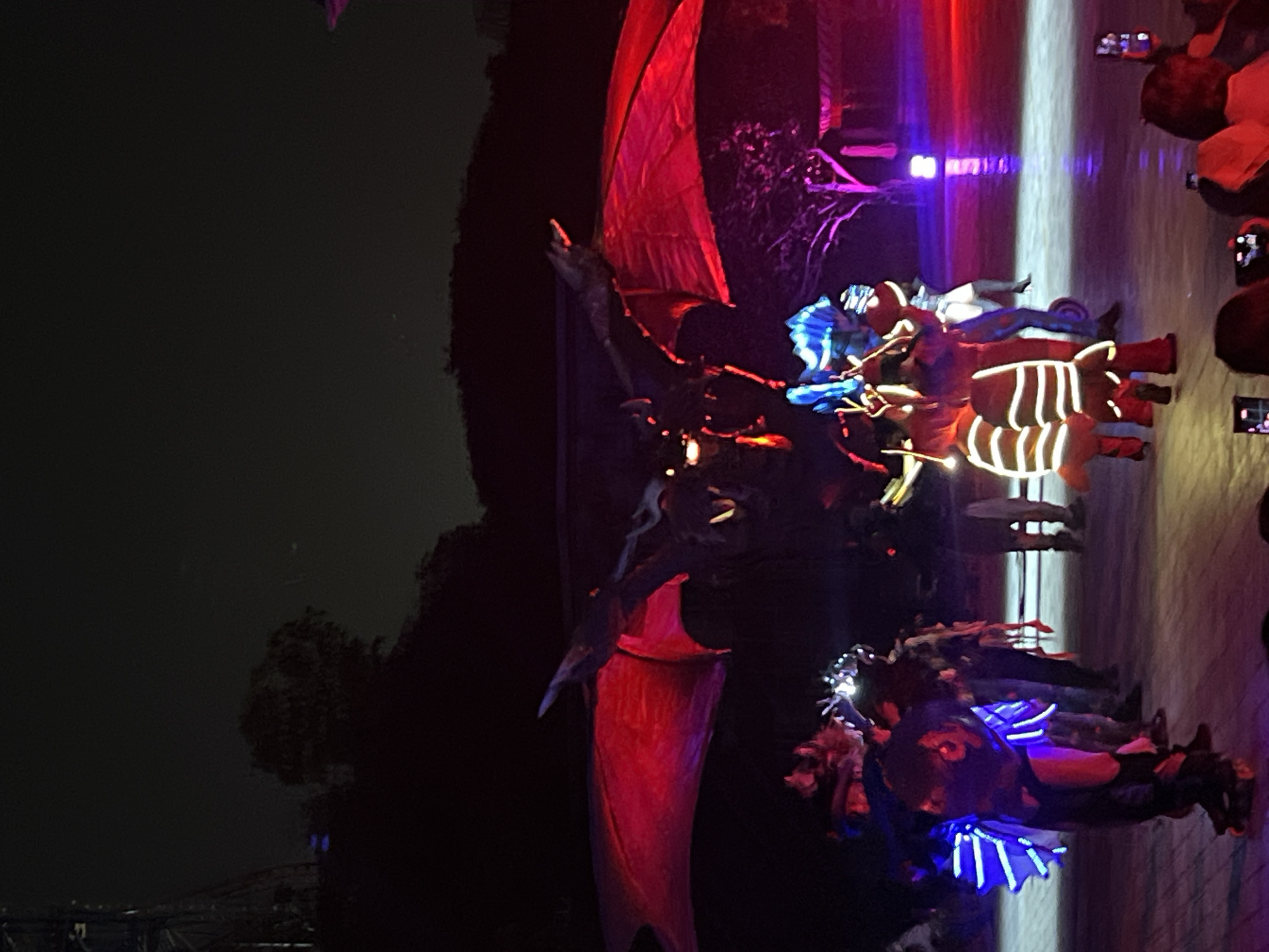 The image shows a performance in Nha Trang, Vietnam. A large red structure with protruding branches is visible to the left. To the right, there are people in costumes with lights. Some of the costumes are red with white lights, and others are blue with white lights. In the background, there is a light-colored wall with vertical streaks of red and blue light. There are also spectators in the background, some of whom are recording the performance with their phones.