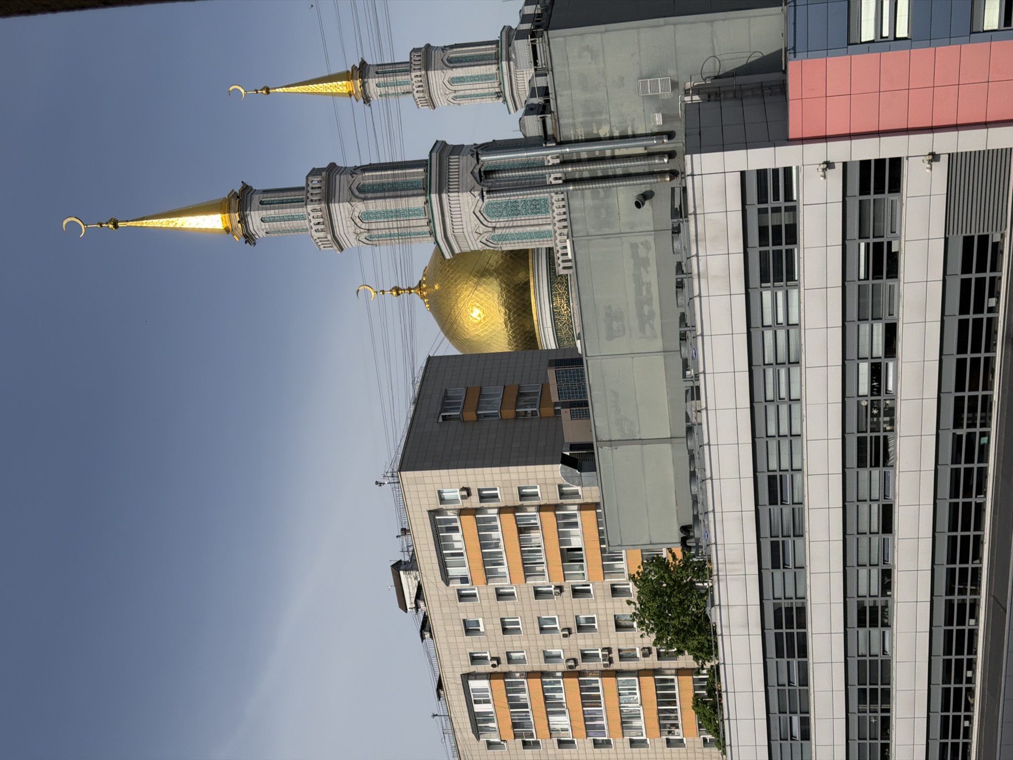 The image shows the Moscow Cathedral Mosque located in Moscow, Russia. The mosque features a large golden dome and two tall minarets topped with golden crescents. Modern buildings with multiple windows and pink accent panels surround the mosque. Overhead power lines are visible against the blue sky.
