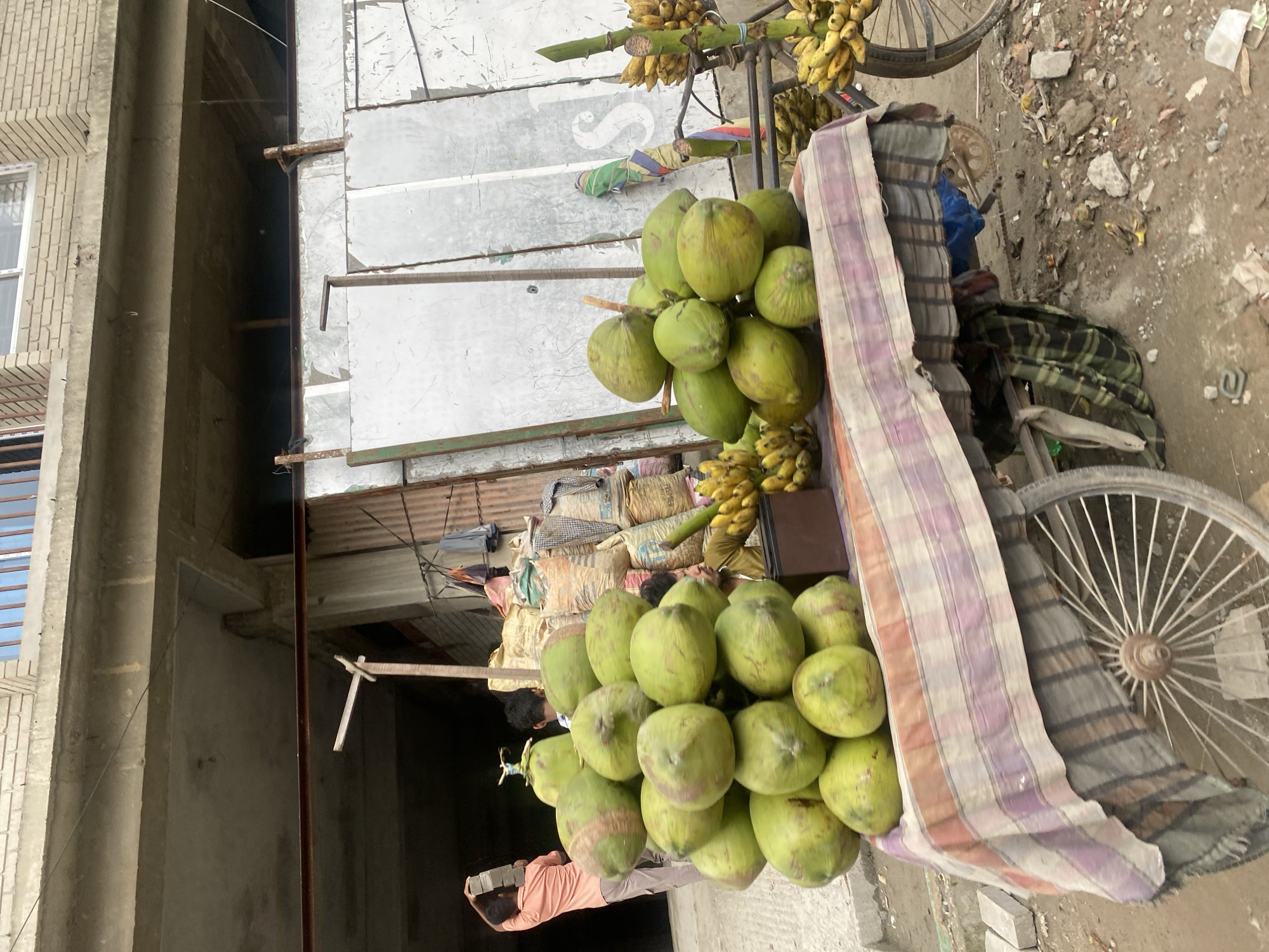 The image depicts a street vendor's cart laden with green coconuts and bananas, parked on a dirt and rubble-strewn street in Dhaka, Bangladesh. The cart, draped with a colorful, striped cloth, has a large spoked wheel visible on its left side. Behind the cart, stacks of large, woven sacks, likely containing cement or grains, are piled high against a corrugated metal structure. A man in an orange shirt is seen in the background carrying a stack of grey bricks. To the right of the cart, bunches of yellow bananas hang from a metal stand. The overall environment suggests a busy, outdoor marketplace.