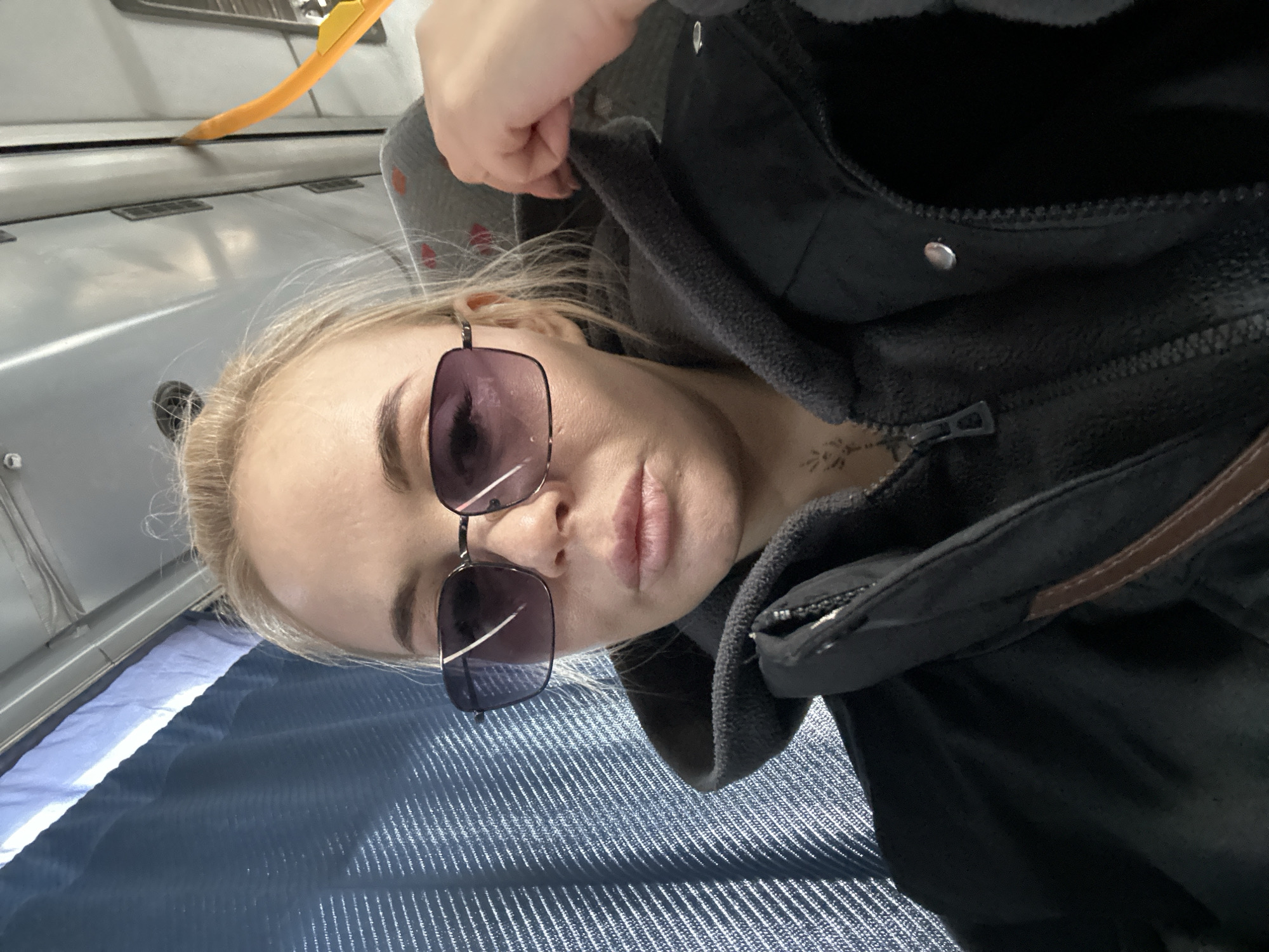 A fair-skinned person with blonde hair is shown in a selfie-style close-up shot, tilted slightly to the side. The person is wearing sunglasses with dark-tinted lenses and a dark jacket with a hood. A grey hooded sweatshirt is visible underneath the jacket. A small tattoo is visible near the person's neck. The background consists of blue fabric with a textured pattern, and the interior of what appears to be a bus or train, based on the visible overhead compartment. Based on the prompt, the location is Megion, Russia.