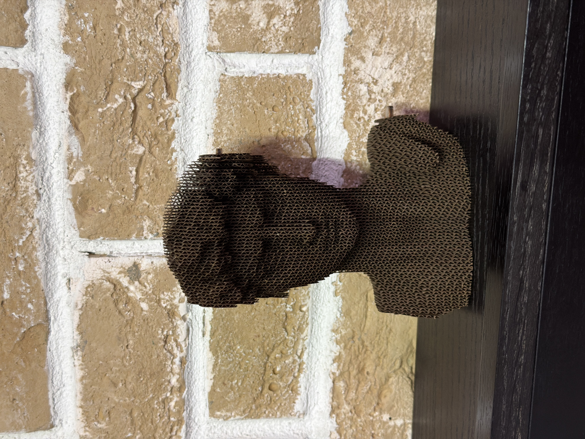 A low-angle, close-up shot reveals a brick wall adjacent to a dark-colored wooden door frame, on which a cardboard sculpture of a camera is mounted. The brick wall exhibits a textured, multi-toned beige color with white grout highlighting the brick pattern. The cardboard camera is composed of multiple layers, creating a three-dimensional form. The door frame, located on the right side of the image, is made of dark wood with visible grain patterns. The setting is possibly an interior space, and there are no visible people or indications of location relative to Moscow, Russia.