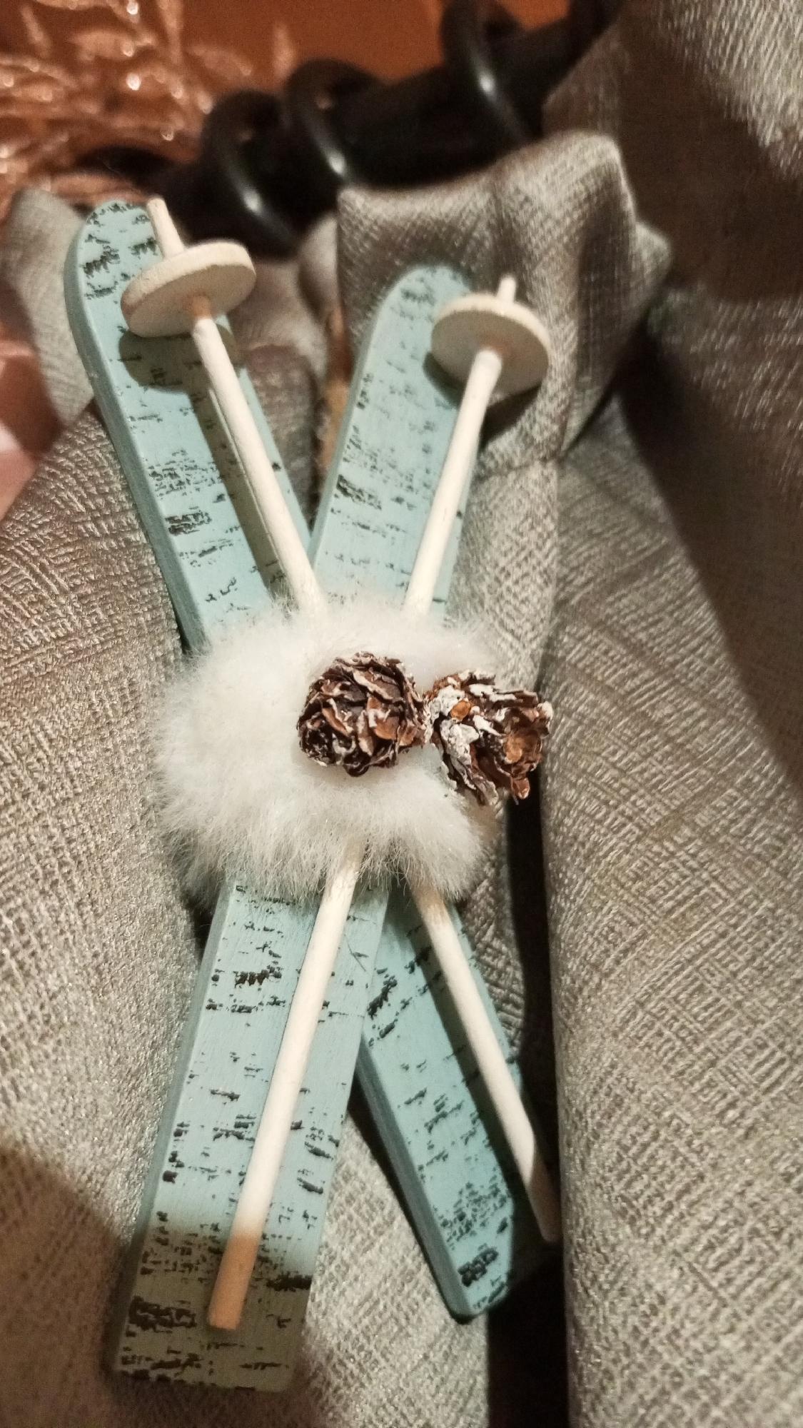 The image shows a pair of miniature crossed skis with poles, tied together at the center with a white, fuzzy pom-pom and two small pinecones. The skis are light blue with distressed black markings. The poles are white. The skis are resting on a textured, gray fabric, possibly a blanket or upholstery. The background includes a dark, out-of-focus object and a glimpse of a shiny, pinkish-gold surface. No people are visible. The location in the city of Юдино, Russia cannot be confirmed from this image.