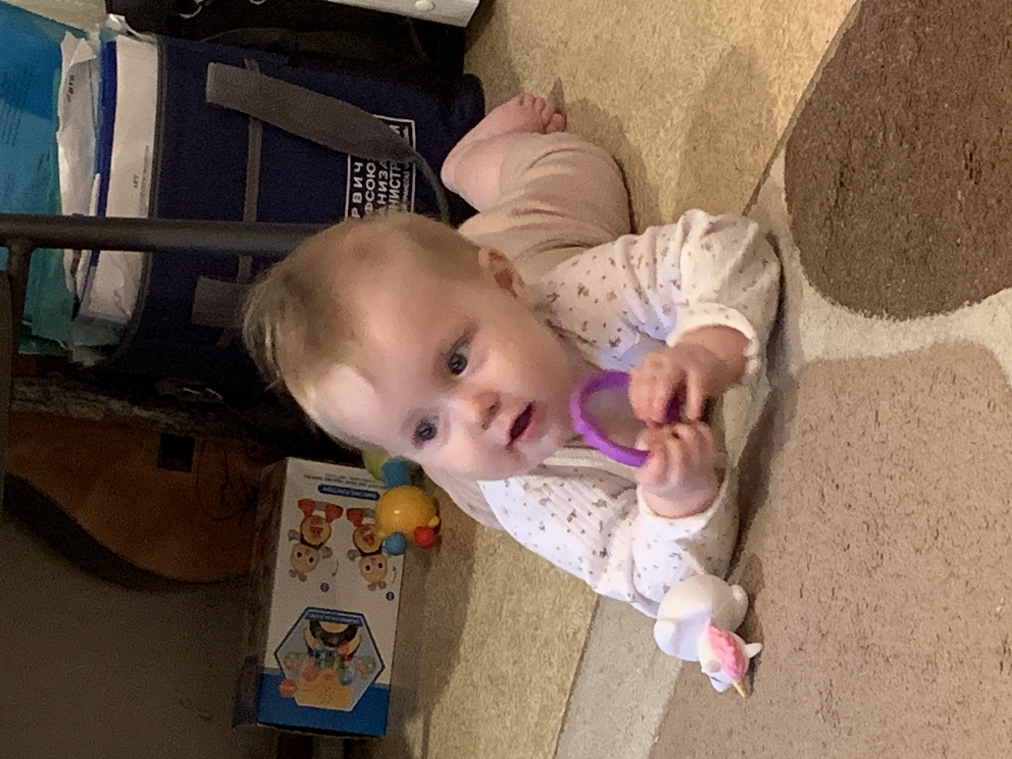 A baby lies on a carpet, partially tan with a brown accent. The baby wears a long-sleeved white shirt with small patterns and light pink pants. The baby holds a purple ring-shaped toy. A box with cartoon bees and a yellow ball with colored pegs sit nearby. The location is implied to be in the city of Большой Букор, Russia, based on external information (not visible in the image). Other objects include a black bag, what appears to be a guitar case, and a unicorn toy. The baby is looking at the camera.
