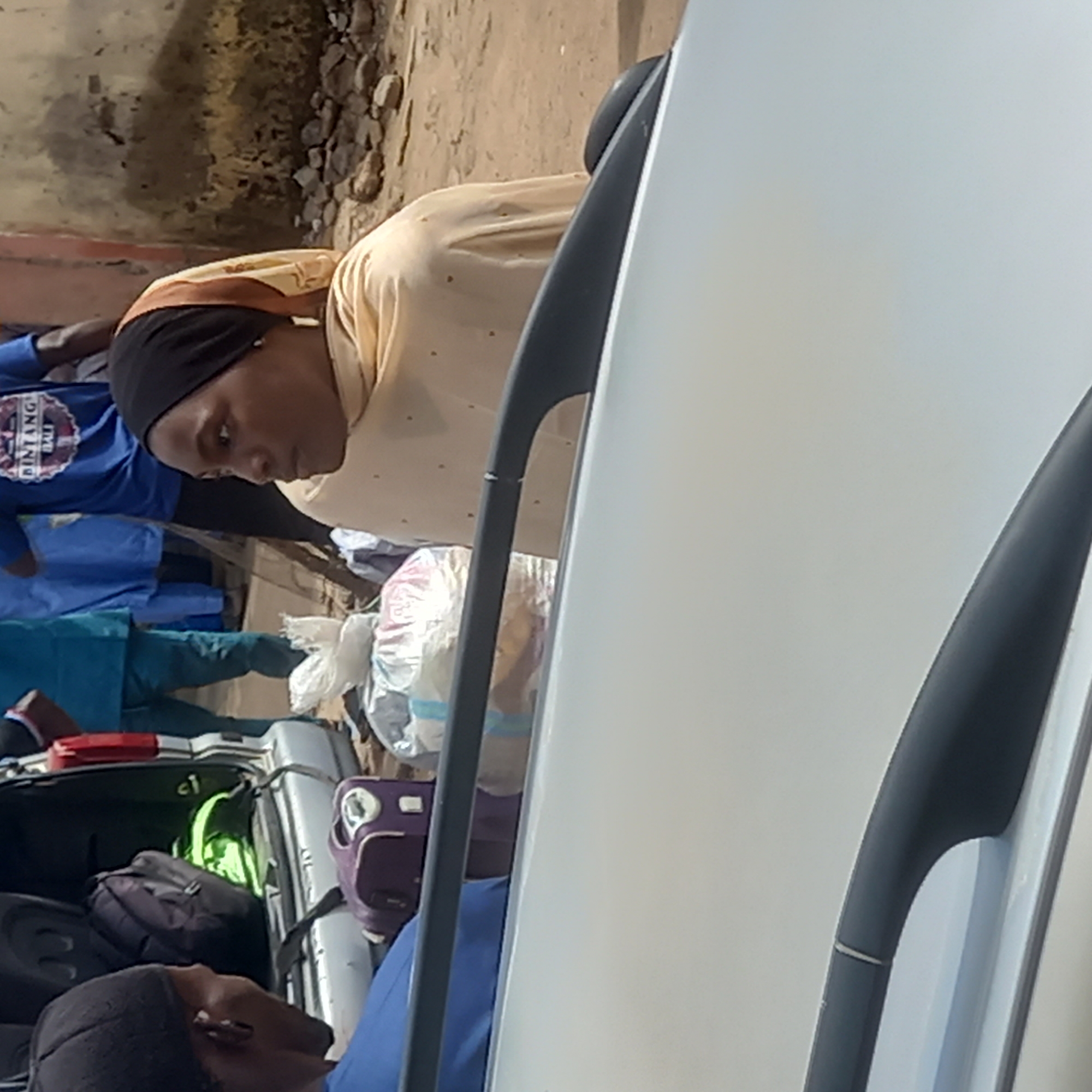 The image depicts a scene likely in Dadi Riba, Nigeria, with a parked white car partially visible on the right. A woman wearing a black head covering and light yellow garment is looking down. Behind her, individuals are visible, one wearing a blue shirt with "BINTANG BALI" printed on it. Luggage, including suitcases and bags, is packed into the trunk of a silver car. The surrounding environment suggests an outdoor location.