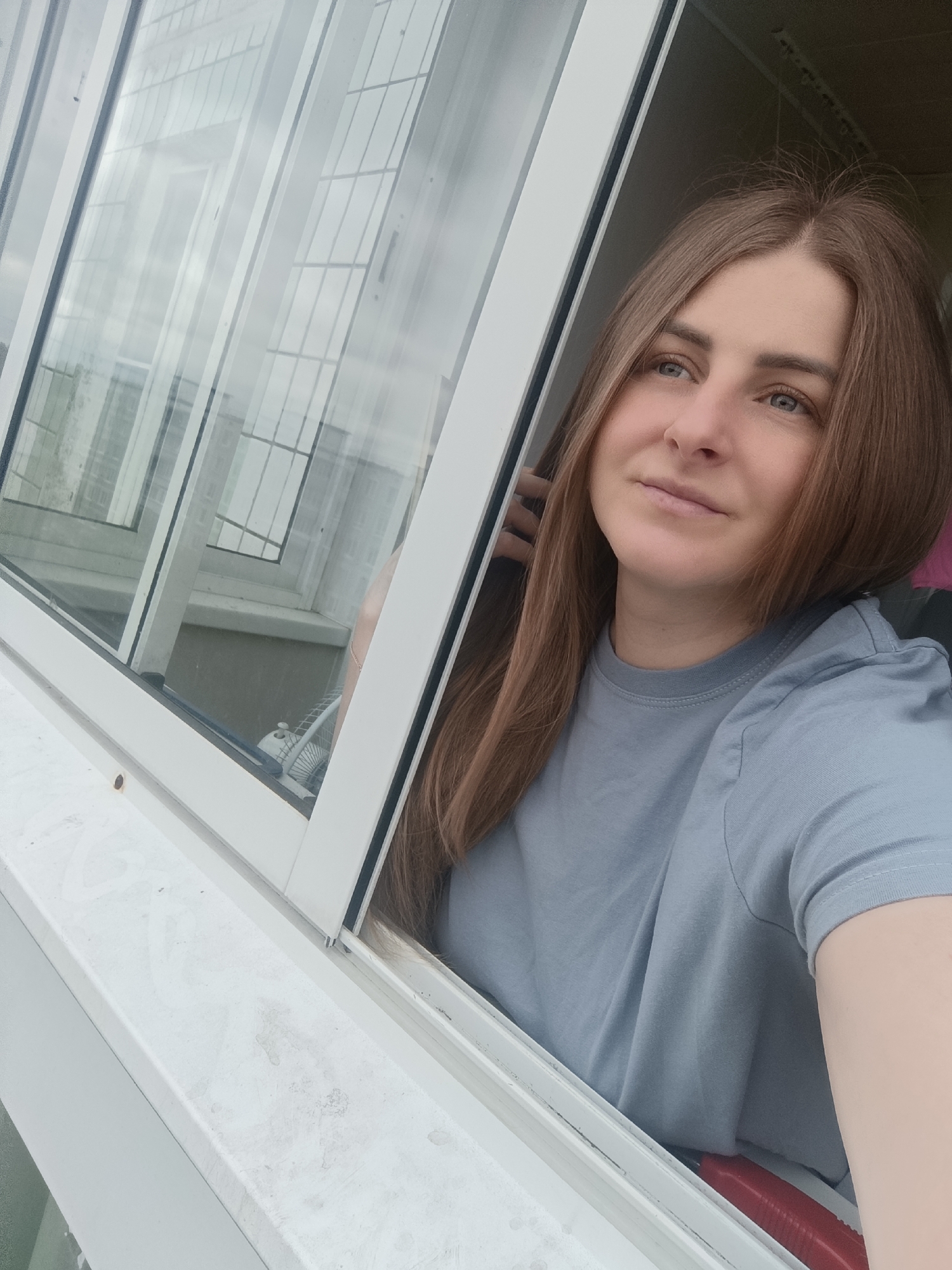 A woman with long brown hair is looking out of a window. The window is white and reflects the building across the street. The woman is wearing a light blue t-shirt. A fan is visible inside the window. The location is Unknown, Unknown.