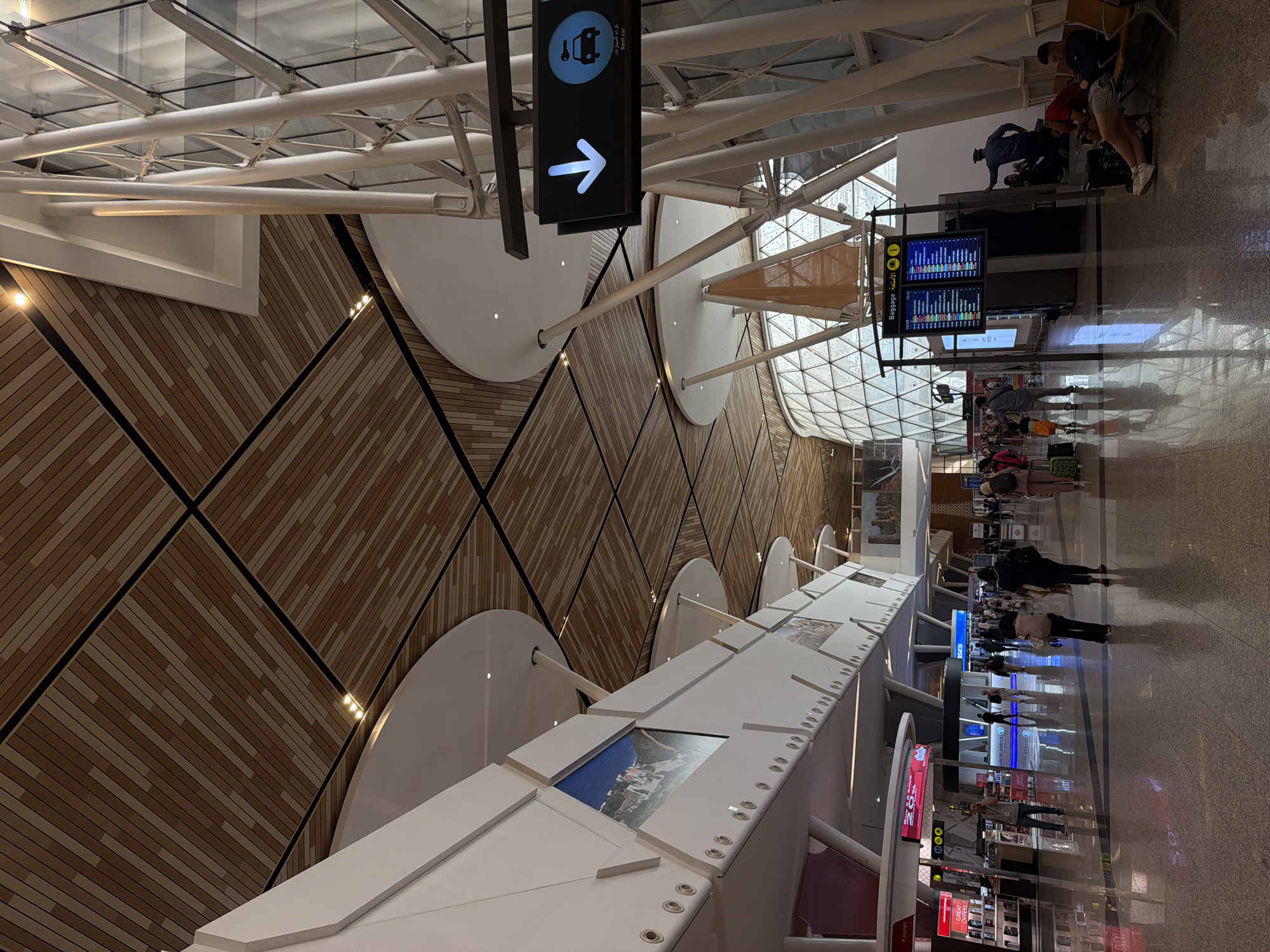 This is an interior photograph of a modern airport terminal, likely Marrakech Menara Airport (RAK) in Marrakesh, Morocco, given the architectural style. The scene depicts a bustling concourse with travelers moving about. The ceiling is a prominent architectural feature, constructed with wooden slats arranged in a geometric pattern, supported by large white structural columns. Large windows, framed by metallic structures, allow natural light to enter.
In the foreground, the polished floor reflects the overhead lighting and the activity within the terminal. Several individuals are walking, some with luggage. On the left side of the image, there are retail outlets, including one with a red sign indicating "DUTRY" and "GREAT OFFERS". Further down the concourse, a "Baggage" information board is visible, displaying flight information in multiple languages. To the right of the baggage claim, people are seated on benches, some looking at their phones, while others rest. A blue directional sign with a white arrow pointing down and an icon of a car and key, labeled "Rent car", is affixed to a white pillar. In the background, a large, geodesic dome-like structure made of glass and metal is partially visible, contributing to the futuristic aesthetic of the airport. The overall impression is one of a clean, spacious, and contemporary transportation hub.