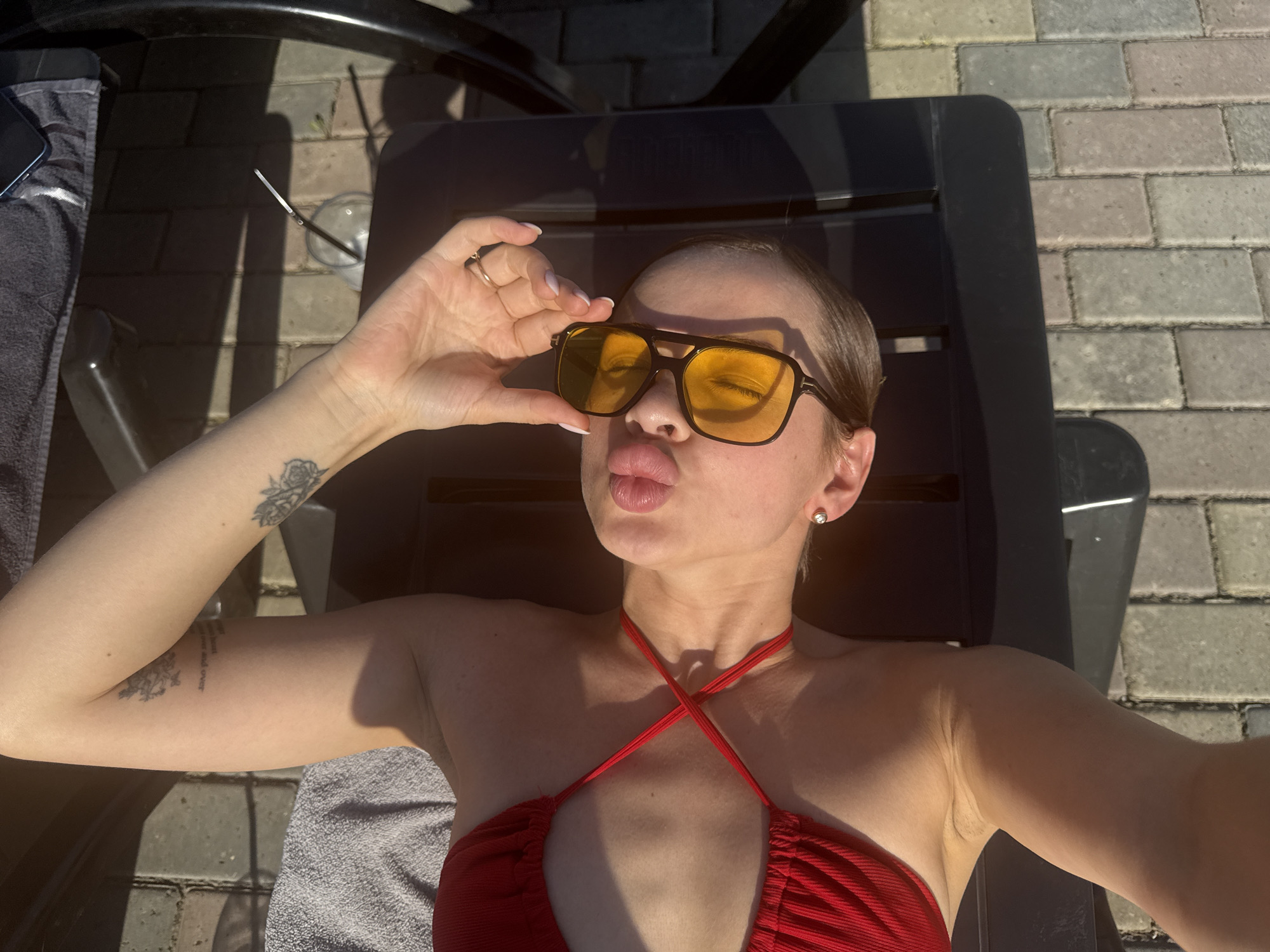 A fair-skinned woman with short, light-colored hair is reclining on a black plastic lawn chair outdoors. She is wearing a red swimsuit with crisscrossing straps at the neckline and yellow-tinted sunglasses. She is puckering her lips and holding the sunglasses, partially obscuring her eyes. Tattoos are visible on her left arm. A glass with a straw is next to the chair. The background consists of gray pavers and other lawn chairs. The image appears to be taken in direct sunlight. It cannot be proven that the location of this image is in Гамово, Russia.