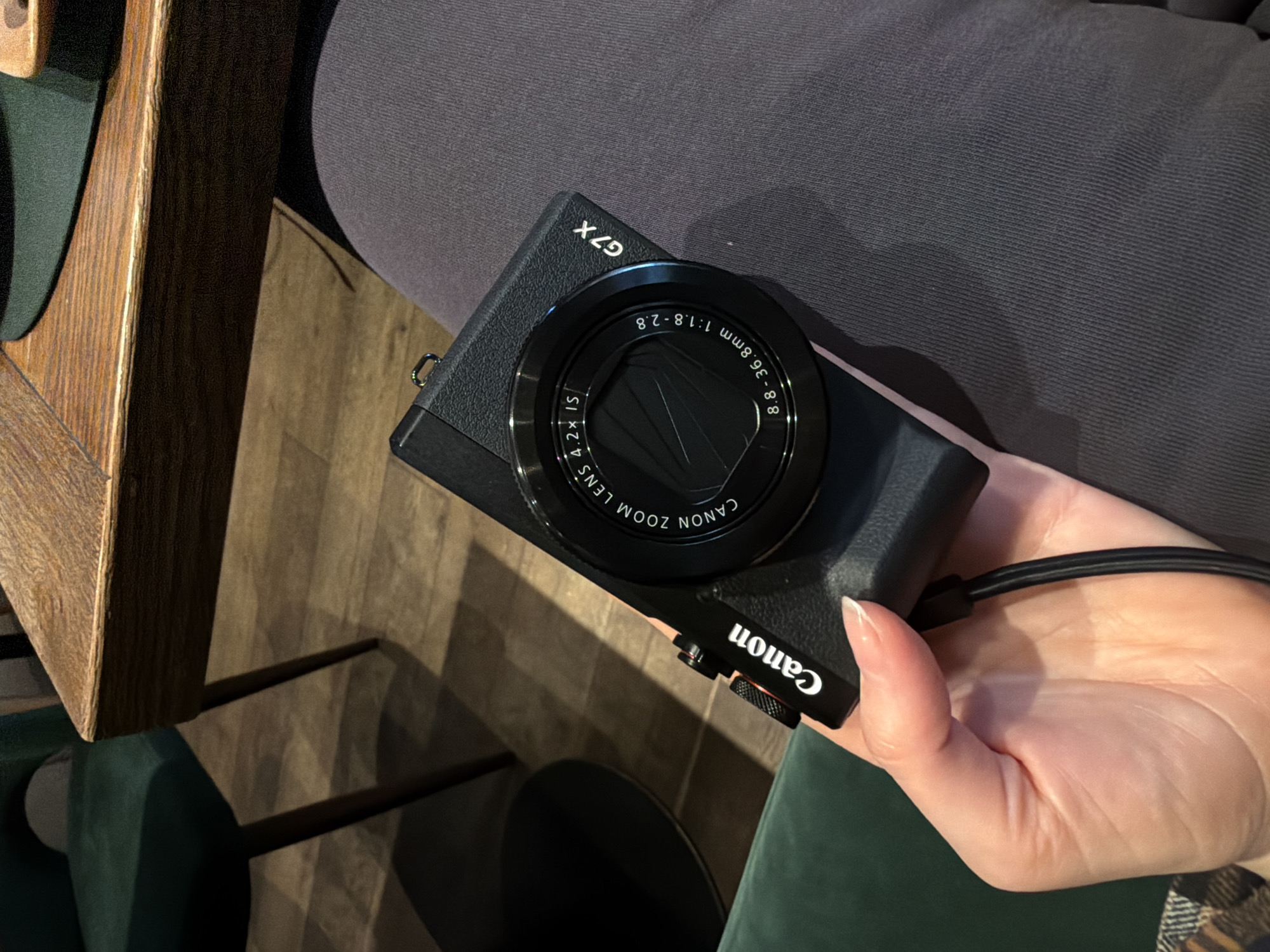 The image shows a Canon G7X digital camera held in a person's hand. The camera is black and has a prominent lens with markings indicating a focal length range of 8.8-36.8mm and an aperture of f/1.8-2.8. The camera's model, "G7X," is visible on the top surface. The hand holding the camera appears to be fair-skinned. The camera strap is black and attached to the side. The background includes a grey surface, possibly clothing, a portion of a wooden table or furniture with a dark stain, and a glimpse of green upholstery. There's no information in the image to suggest it's in Moscow, Russia.