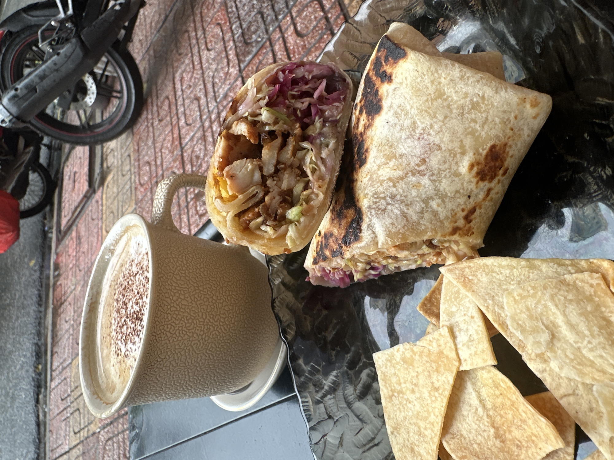 A still life shows a half-eaten wrap cut in half to show the ingredients of chicken, shredded lettuce, and purple cabbage, a triangular folded wrap, and a pile of triangular tortilla chips. Next to this is a coffee cup filled with a latte with sprinkles. The picture is taken at a low angle, and in the background is a motorcycle and a textured brick ground. This photo was taken in Nha Trang Ward, Vietnam.
