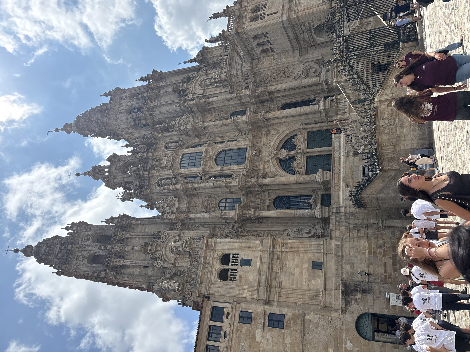 The image shows a low-angle view of the facade of the Cathedral of Santiago de Compostela in Spain, with a group of people gathered in front of it. The cathedral is a large, ornate stone structure with intricate carvings and multiple spires. The sky above is blue with scattered white clouds. In the foreground, a crowd of people, many wearing white t-shirts, are milling about on a cobblestone plaza. Some are looking at the cathedral, while others are talking to each other or taking pictures. The scene appears to be a sunny day, and the atmosphere is one of tourism and admiration of the historical landmark.