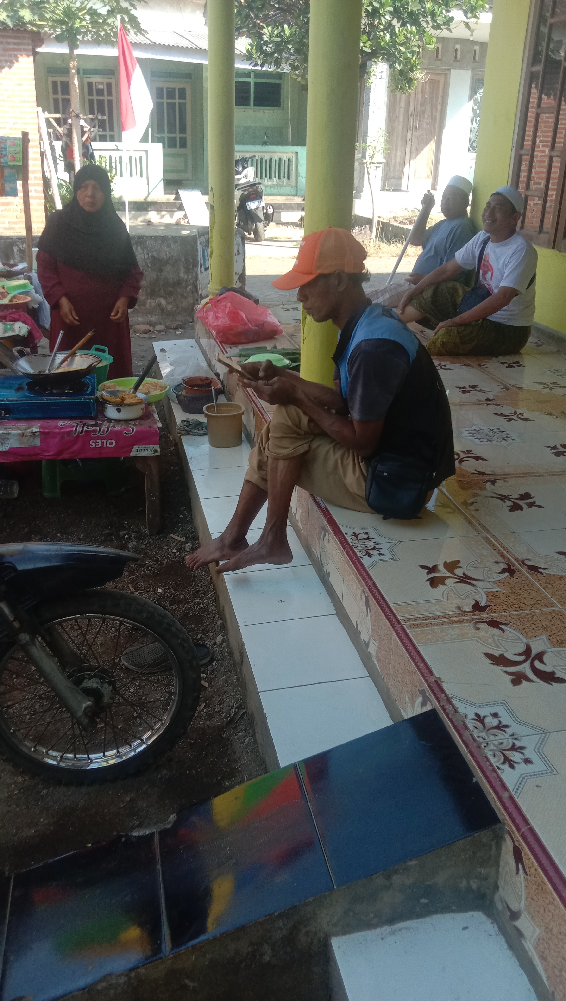 In Ngempit, Indonesia, an outdoor scene unfolds on a tiled patio adjacent to a building with a veranda supported by yellow columns. A woman wearing a dark hijab and a maroon dress stands near a makeshift food stall, which includes a portable stove and various containers. To her right, a man in an orange cap and a blue and black shirt sits on the edge of the patio, looking down at a mobile phone he holds in his hands. His legs are bare, and he wears tan pants. Further to the right, two other men are seated on the patio. One wears a white shirt and a white skullcap, and the other wears a blue shirt and a white skullcap. Both men are looking in the same direction as the man with the phone. A red and white Indonesian flag is visible in the background, attached to a pole in front of the building. A motorcycle is parked on the left side of the image, partially obscuring the lower section of the food stall. The environment appears to be a residential area, possibly during a community gathering or local market.