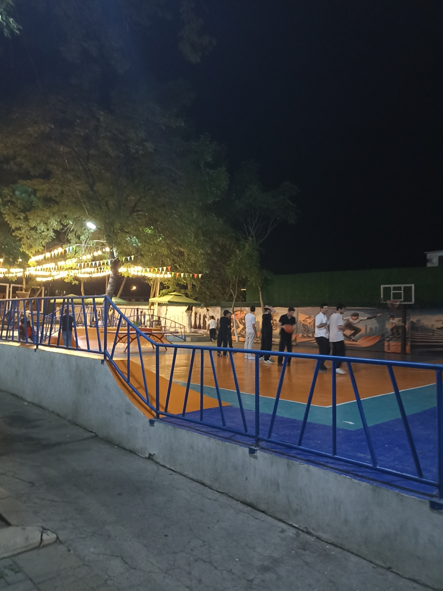 The image depicts an outdoor basketball court at night. Several individuals, appearing to be young men, are present on the court, some of whom are engaged in playing basketball. The court itself has a colorful surface with orange and blue sections, marked with white lines. A basketball hoop with a backboard is visible in the background. The area is illuminated by artificial lighting, including string lights decorated with small flags draped over trees and poles. Behind the court, there's a green fence or wall with a mural depicting figures in action. A blue metal railing separates the court from a slightly elevated walkway or seating area. The overall environment suggests a recreational area, possibly a park or a sports facility, in Gusar, Azerbaijan.