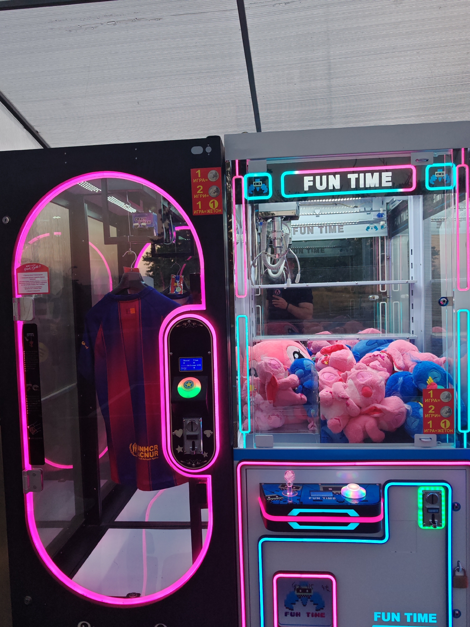 This is an eye-level, medium indoor shot of two arcade claw machines side-by-side. The machine on the left, outlined with pink neon lights, contains a Barcelona soccer jersey and other smaller items hanging inside. The machine on the right, lit with cyan and pink neon, is full of pink and blue plush toys. Both machines have a "FUN TIME" logo. The images on the machines are written in both English and a Cyrillic language, and one of the machine has "UNHCR ACNUR The UN Refugee Agency" written on the displayed item. The picture was likely taken in Nesebar, Bulgaria based on the Cyrillic text on the machines.