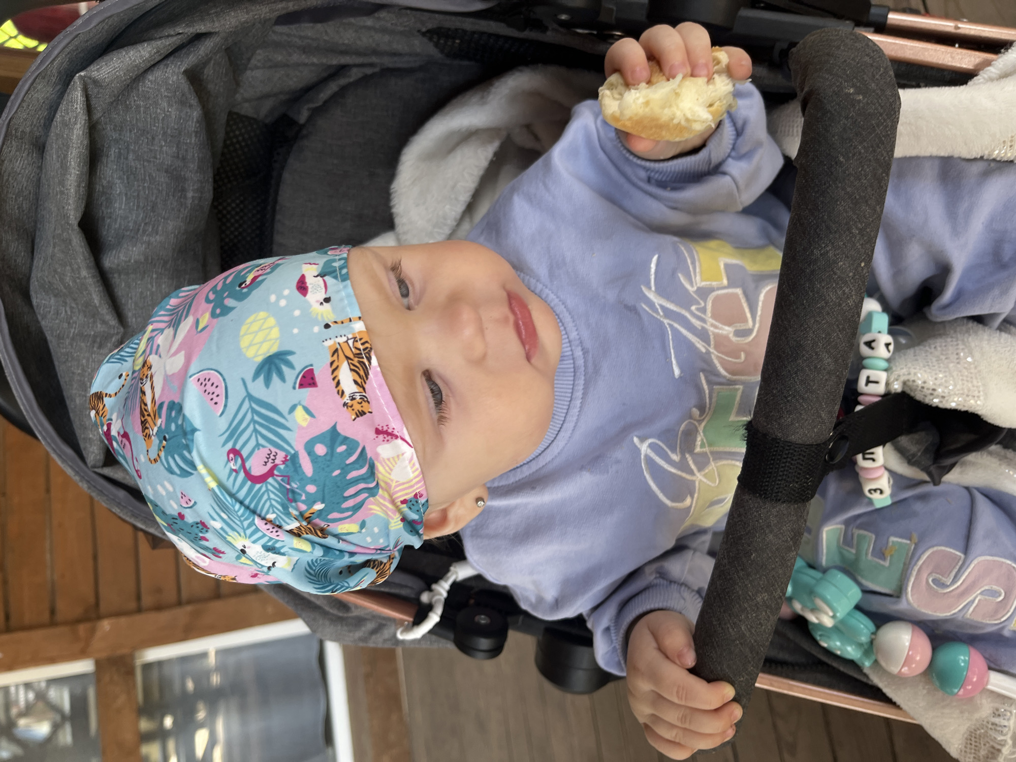 A young Caucasian baby with blonde hair and blue eyes is sitting in a stroller, wearing a light blue sweatshirt and a colorful bandana with a tropical print. The baby is holding a piece of bread in its right hand and is looking down. The stroller is gray and has a dark gray handlebar. Attached to the handlebar are several colorful baby toys, including a rattle. The background shows a wooden structure, possibly a porch or deck, with wooden planks. The lighting suggests it is daytime. The location is the city of Фоки, Russia.
