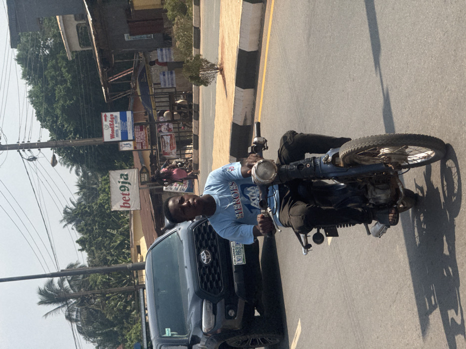 A young male operates a blue-colored, older model motorbike on an asphalt road in Aguata, Nigeria. He wears a light blue t-shirt with "HEROES 10 ATM" print, black cargo pants, and dark sandals, looking slightly to his right. His hands grip the handlebars, and his feet rest on the motorbike's frame. The motorbike features a circular headlight, spoked wheels, and exposed engine components. Behind him and to his left, a dark-colored Toyota Tacoma pickup truck with license plate "LND-863JV" is partially visible. The road has a solid white lane line on the left. To the right of the motorbike is a raised curb, painted with black and white stripes, separating the road from a dirt shoulder. A small green shrub is planted on this median. In the background, across the road, various structures are present, including a white billboard displaying "bet9ja" and "BEN GIG STREET," and a blue sign for "WoodEdge HOTEL & SUITES." Overhead utility poles and multiple power lines crisscross the scene. Several individuals are visible in the distance near the structures, including one person in a pink garment. Dense tropical vegetation, including palm trees, rises behind the buildings. Long shadows are cast on the road, indicating strong sunlight.