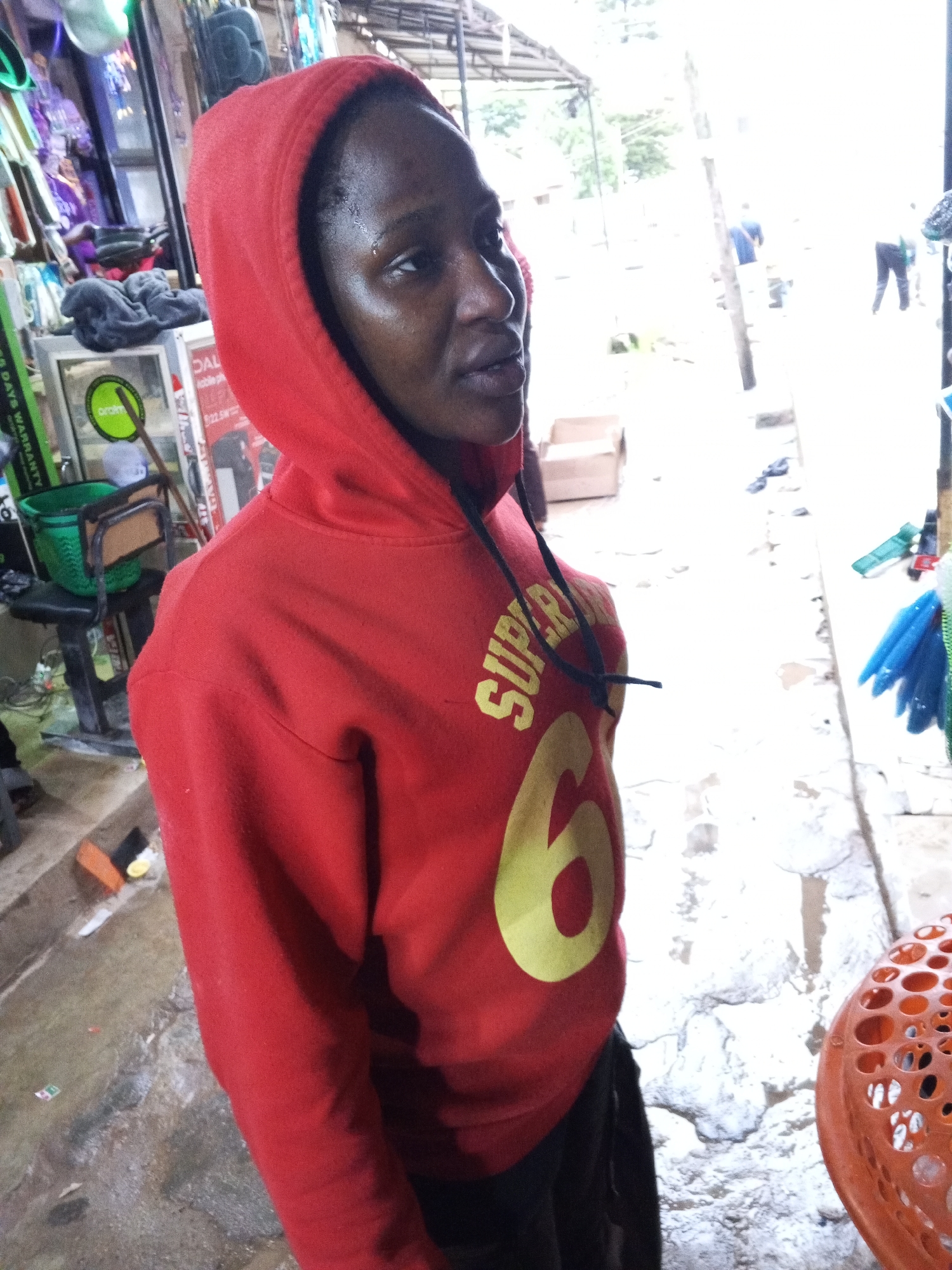 The image shows a young Black woman wearing a red hooded sweatshirt with yellow lettering. She is standing outdoors in what appears to be a marketplace in Jos, Nigeria. The ground is wet and appears to be concrete with some debris scattered on it. In the background, there are stalls with various items for sale, including what looks like electronics and other merchandise. There are also other people visible in the distance, some walking and others standing. The lighting suggests it is daytime, possibly overcast or with dappled sunlight.