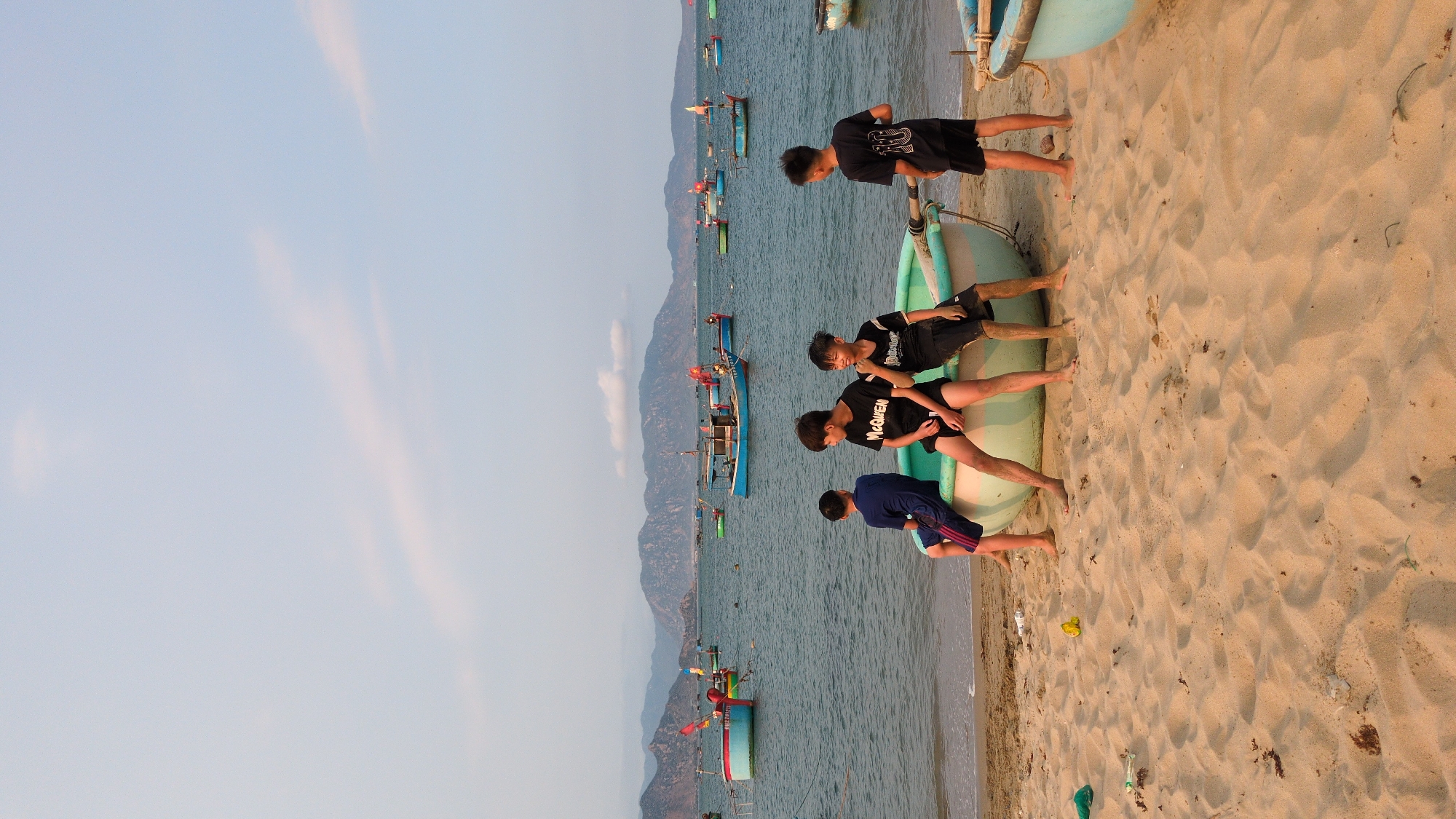 Captured in Phan Rang – Tháp Chàm, Vietnam, the photograph depicts a beach scene. Four young males are interacting with a teal boat on the sandy shore. One stands beside the boat, while the other three are seated on it. The sea, dotted with numerous other boats, stretches out to the mountainous horizon. The sky is clear, with a slight gradient in color. The beach sand is a light tan color. Debris is scattered on the beach.