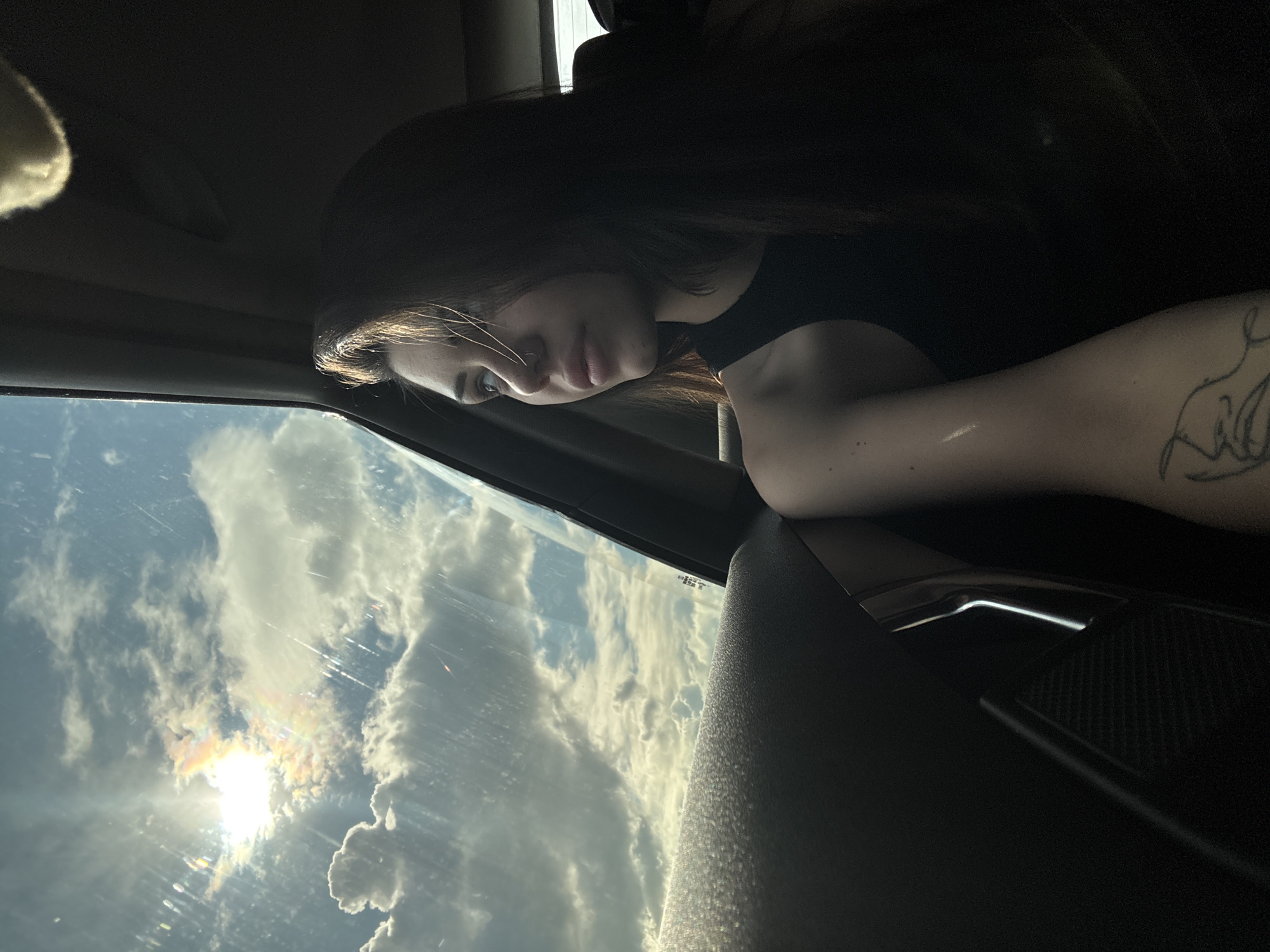 The image captures a moment inside a car in Herakovo, Bulgaria, showcasing a person looking out a window towards a bright, cloudy sky. A young woman, with long dark hair, is positioned in the foreground, her profile visible as she gazes intently out. She wears a dark top, and a tattoo is visible on her right upper arm. Her expression appears calm and thoughtful.
The scene outside the car window reveals a vibrant daytime sky. The sun shines brightly through scattered white clouds, creating a distinct sun dog or iridescence effect with rainbow-like colors around it. The sky is predominantly blue, indicating fair weather. Slight reflections and minor streaks are visible on the car window glass. Elements of the car's interior, including the dark headliner, door panel, and dashboard, frame the view. Faint text, notably "HST," is visible on a sticker adhered to the car window. The overall ambiance suggests a pensive moment during a journey, with an appreciation for the natural beauty of the sky.