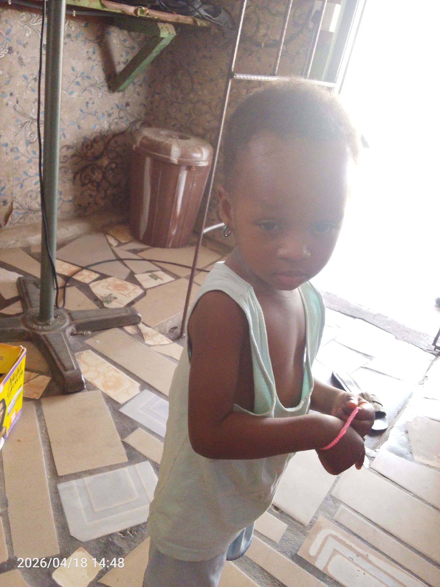 A young child stands indoors. The child is wearing a light-colored sleeveless shirt. The flooring appears to be tiled with a mix of tan and white tiles. A brown plastic bin sits in the background near a wall with patterned wallpaper. A metal stand of some kind is to the left of the child. The time and date stamp "2026/04/18 14:48" is visible in the lower left corner. The setting appears to be an interior space with bright light coming from an unseen window or doorway. Location in the city of Lokoja, Nigeria cannot be confirmed based on image details.