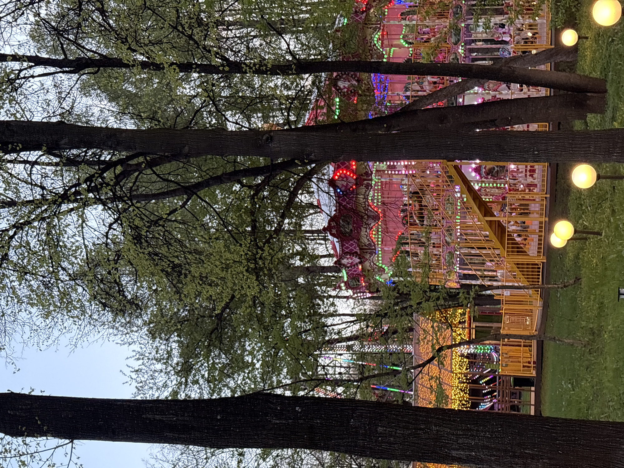 The image depicts an amusement park ride in Perm, Russia, as seen through trees. The ride is brightly lit with various colored lights. Several yellow, globe-shaped streetlights are visible. Trees are in the foreground, obscuring the view of the ride. The sky is visible through the branches. There are no people directly visible.