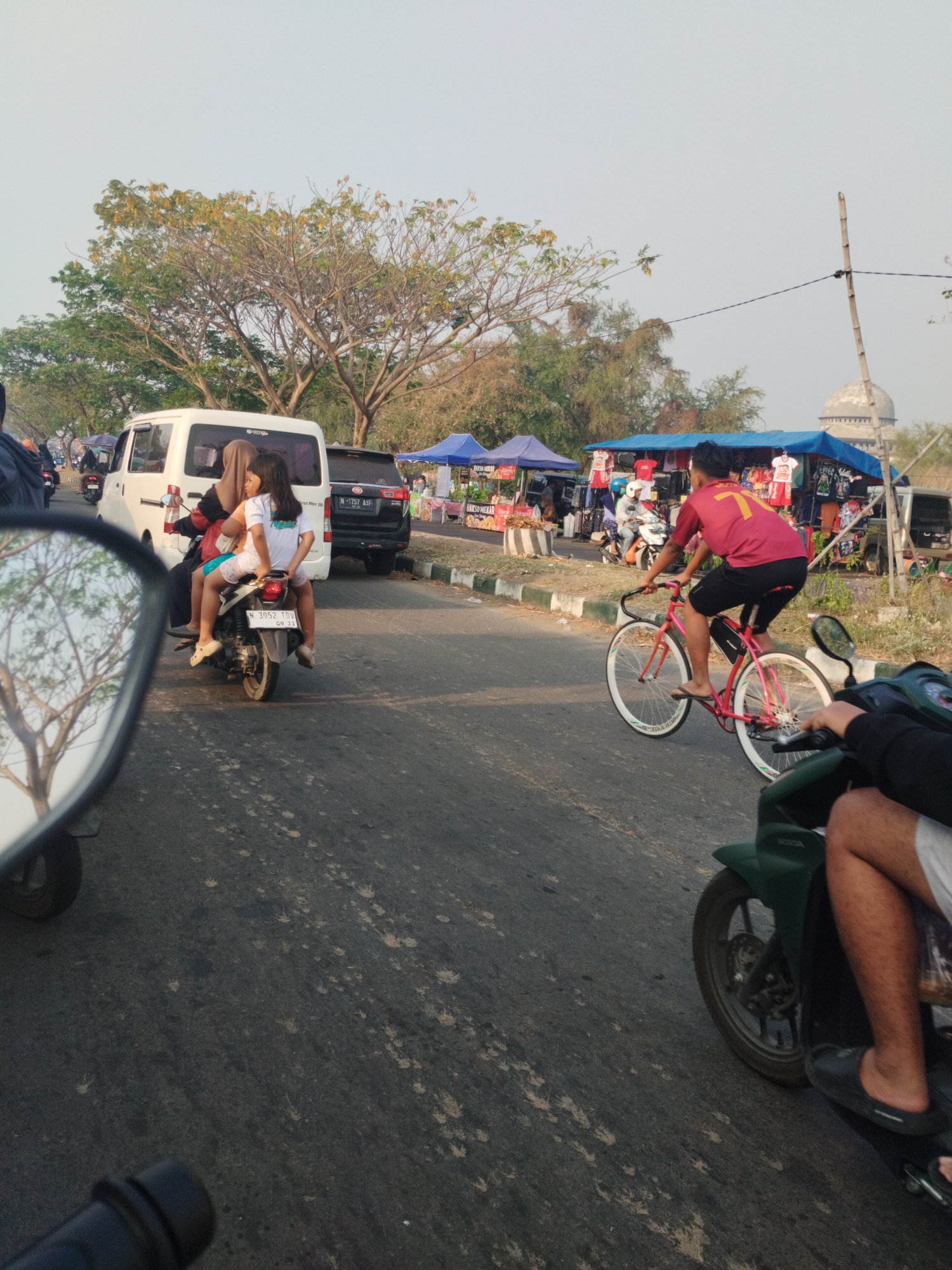 A street scene in Besuki, Indonesia, features a variety of modes of transportation and roadside vendors. In the foreground, a green motorcycle is partially visible on the right, with the rider's legs and feet in view. To the left, the reflection of trees and sky is visible in the mirror of another unseen vehicle. Further down the road, a pink bicycle is being ridden by a young man wearing a red shirt and black shorts. Behind him, a motorbike carries two passengers, a woman in a hijab and a young girl. A white van is parked behind the motorbike. In the background, a row of tents set up as stalls displays clothing for sale, and a dome-shaped structure is visible in the distance. The road appears to be paved and is lined with sparse vegetation. The sky is overcast, suggesting a daytime scene.