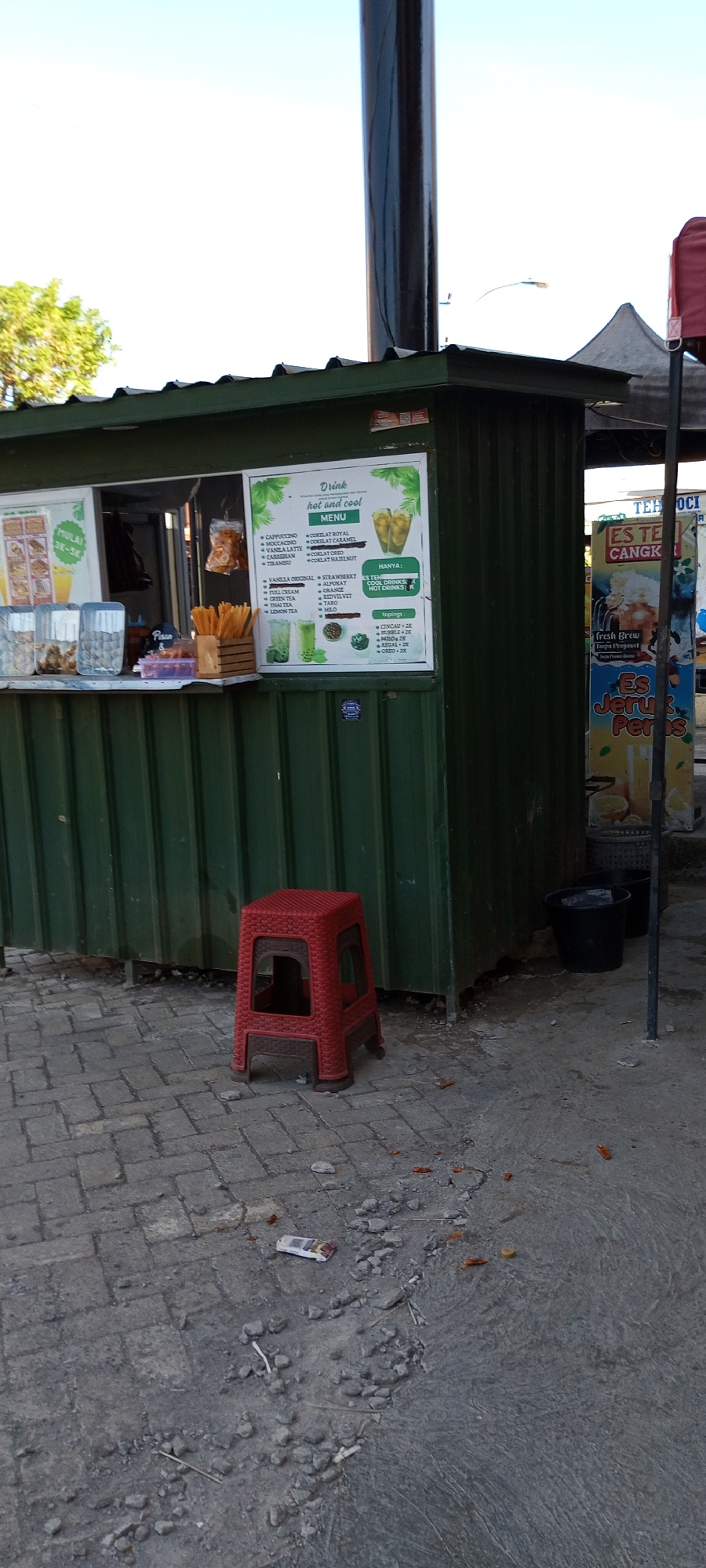 The image depicts an outdoor scene in Assorajang, Indonesia. In the foreground, a red plastic stool sits on a cobblestone surface littered with small stones and debris, including cigarette butts. To the right of the stool, two black buckets are visible.

The central focus is a small, green kiosk with a corrugated metal exterior. A window reveals drinks being served. A sign inside the kiosk displays a menu for "hot and cool" drinks, listing various flavors such as Cappuccino, Mocca, Vanilla Latte, Caramel, Tiramisu, Coklat Royal, Coklat Caramel, Coklat Oreo, and Coklat Hazelnut. Underneath, a section labeled "HANYA:" (meaning "Only:") indicates that "Es Teh" (Iced Tea) and "Cool Drinks" are available for an unspecified price, while "Hot Drinks" are also available. Another section details "topping" options: Cincau, Bubble, Regal, and Oreo, each with an associated price. To the left of the menu, a smaller sign shows prices ranging from "Mulai 3K-5K" (starting from 3,000 to 5,000 Indonesian Rupiah).

Next to the green kiosk, to the right, is a tall, upright sign advertising "ES TEH CANGKIR" (Iced Tea in a Cup), with an image of a person holding a large cup of iced tea. Further to the right, another sign reads "Es Jeruk Peras" (Freshly squeezed orange juice), featuring an image of oranges and a glass of orange juice.

A tall, dark pole stands behind the green kiosk, extending upwards and out of frame, likely supporting some structure or overhead lighting. In the background, a glimpse of green trees and a clear blue sky suggest it is daytime. The overall impression is a street-side beverage stall in a commercial or public area.